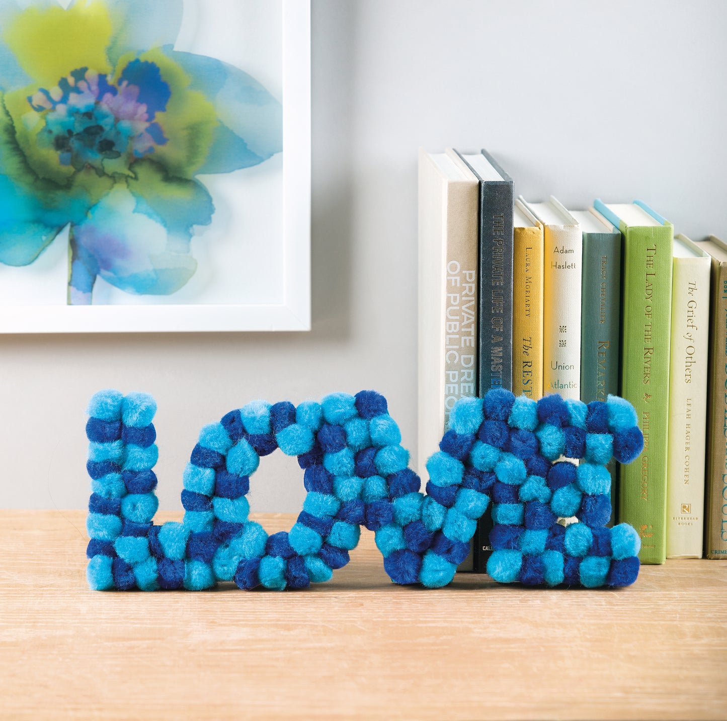 Blue and teal pom-poms from the Pom-Pom Crafts kit by Leisure Arts are arranged to spell LOVE on a wooden surface, displayed beside upright books and underneath a framed floral artwork on a white wall—an easy DIY home décor idea ideal for anyone seeking beginner craft projects with Leisure Arts’ Pom-Pom Crafts.