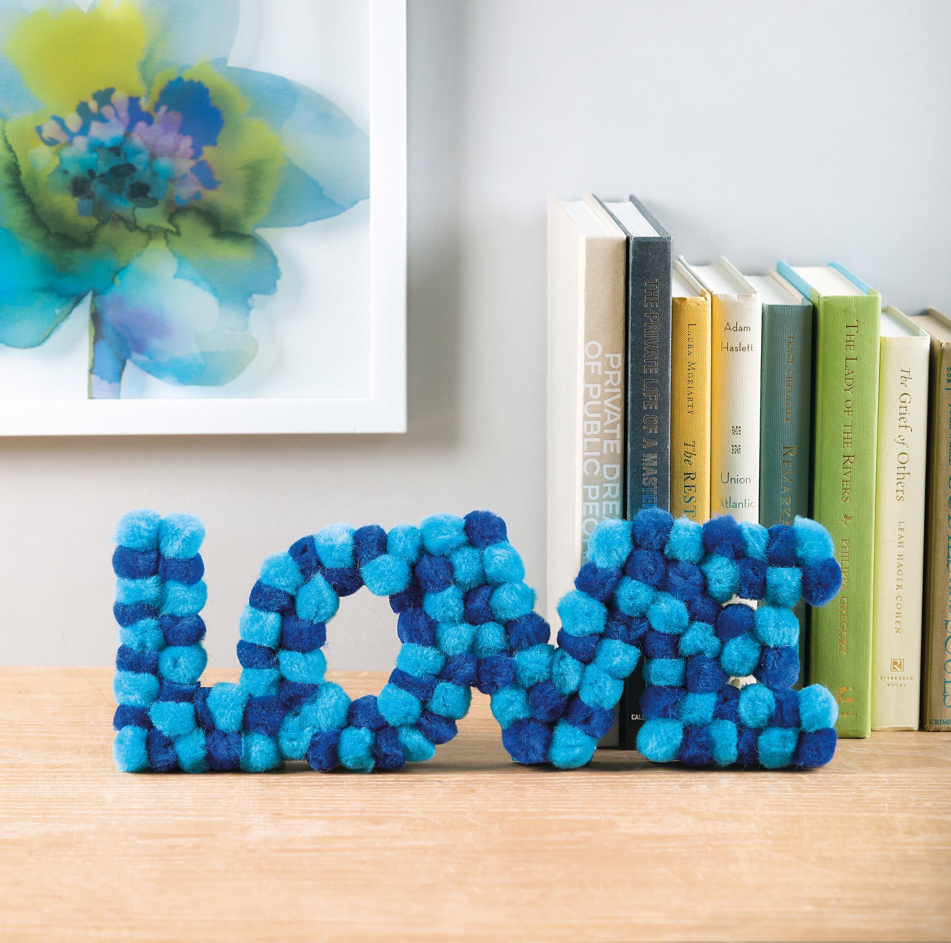 Blue and teal pom-poms from the Pom-Pom Crafts kit by Leisure Arts are arranged to spell LOVE on a wooden surface, displayed beside upright books and underneath a framed floral artwork on a white wall—an easy DIY home décor idea ideal for anyone seeking beginner craft projects with Leisure Arts’ Pom-Pom Crafts.