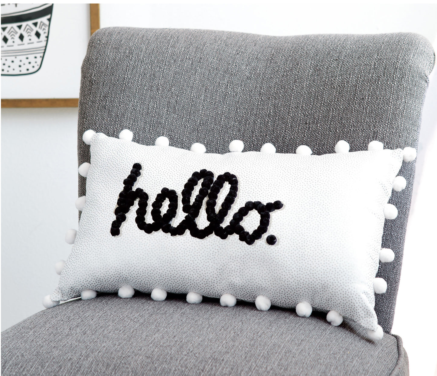 A white rectangular pillow from Leisure Arts’ Pom-Pom Crafts collection, featuring “hello.” written in black cursive and playful white pom-poms along the edges, sits on a gray upholstered chair—perfect for easy DIY home décor inspired by charming pom-pom crafts.