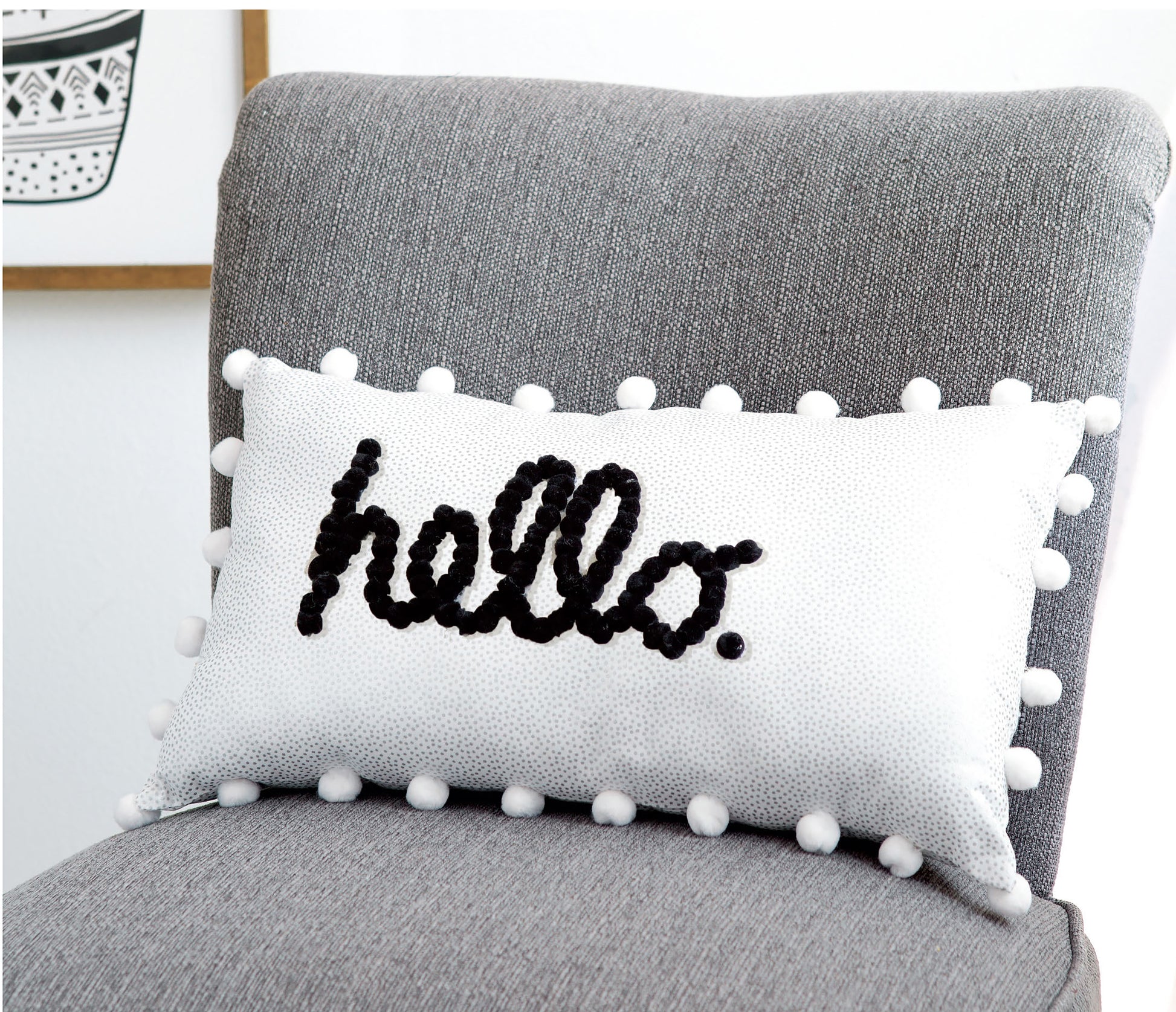 A white rectangular pillow from Leisure Arts’ Pom-Pom Crafts collection, featuring “hello.” written in black cursive and playful white pom-poms along the edges, sits on a gray upholstered chair—perfect for easy DIY home décor inspired by charming pom-pom crafts.