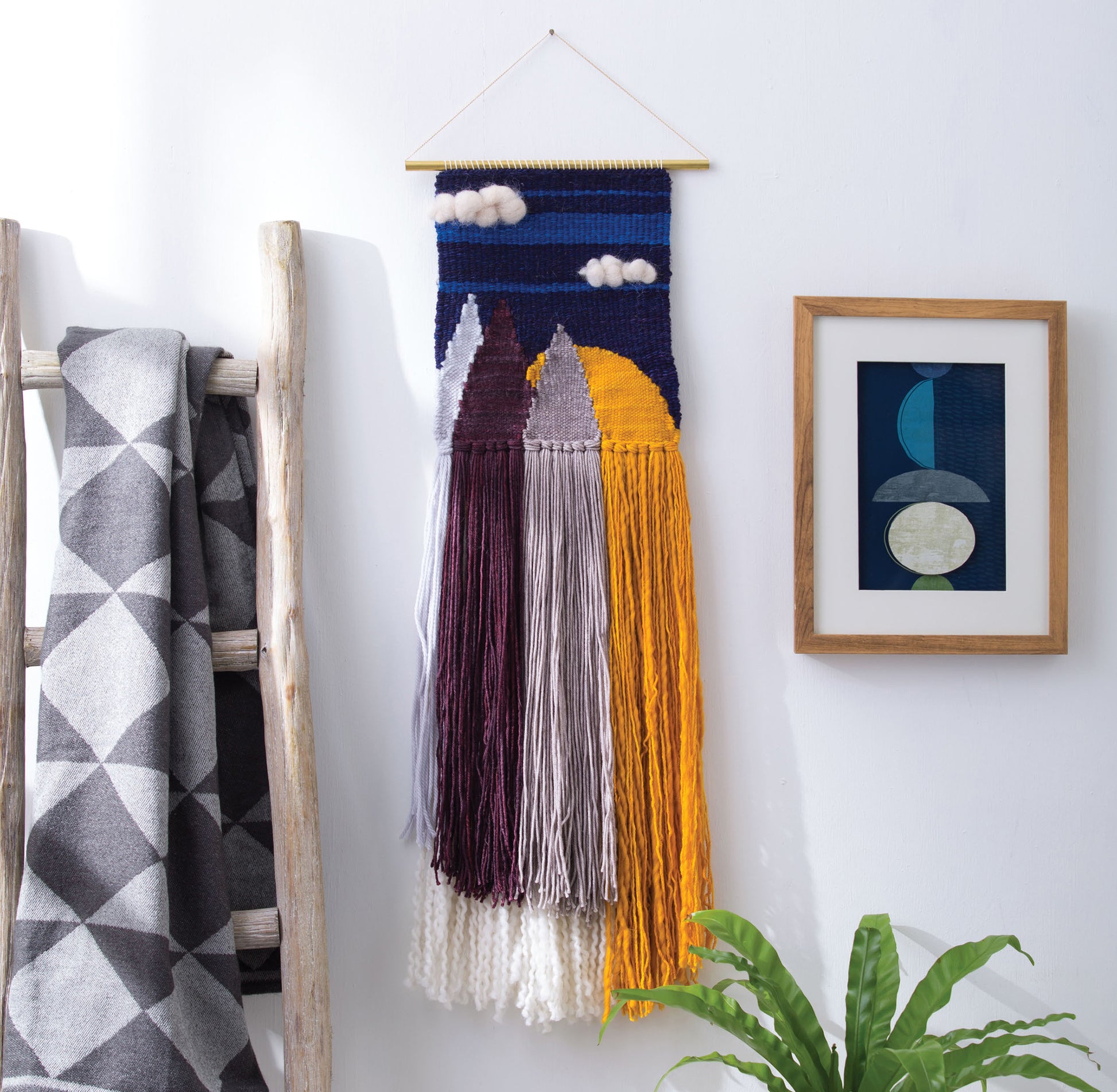 The "Learn to Make Woven Wall Hangings" kit by Leisure Arts features mountain shapes, clouds, and vibrant yarn fringe—perfect for beginners—and is displayed beside a framed abstract print, blanket-ladder, and green potted plant.