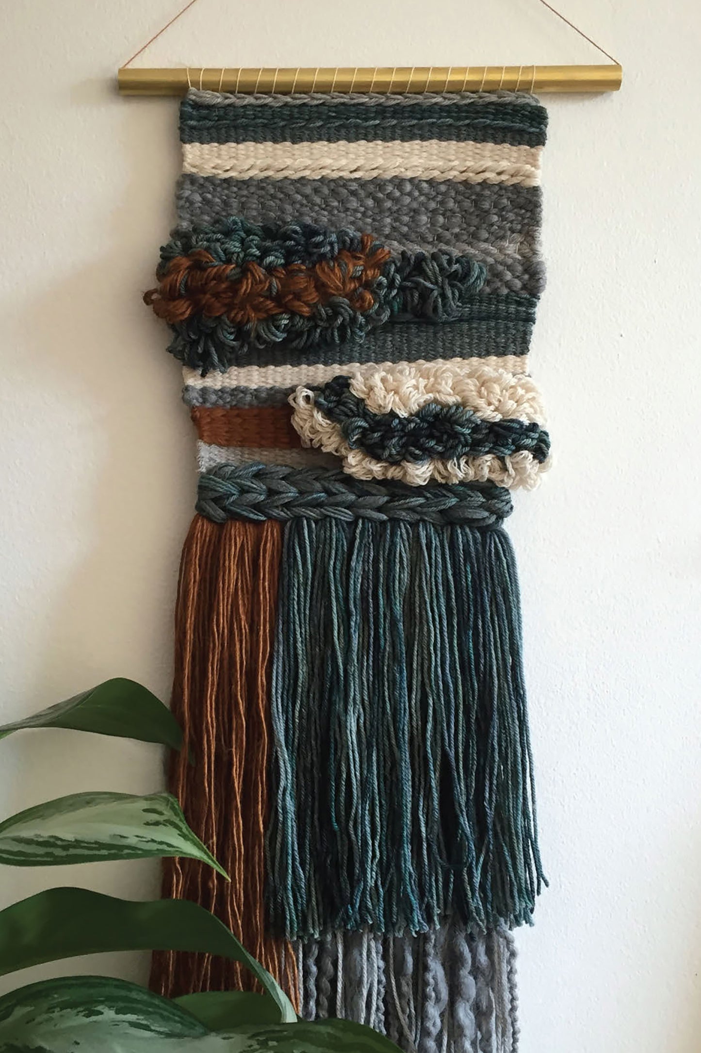 The Leisure Arts "Learn to Make Woven Wall Hangings" kit features textured yarn in green, brown, cream, and gray with braided and looped details, long fringe, a gold top rod, and a plant leaf accent—perfect for frame loom projects.