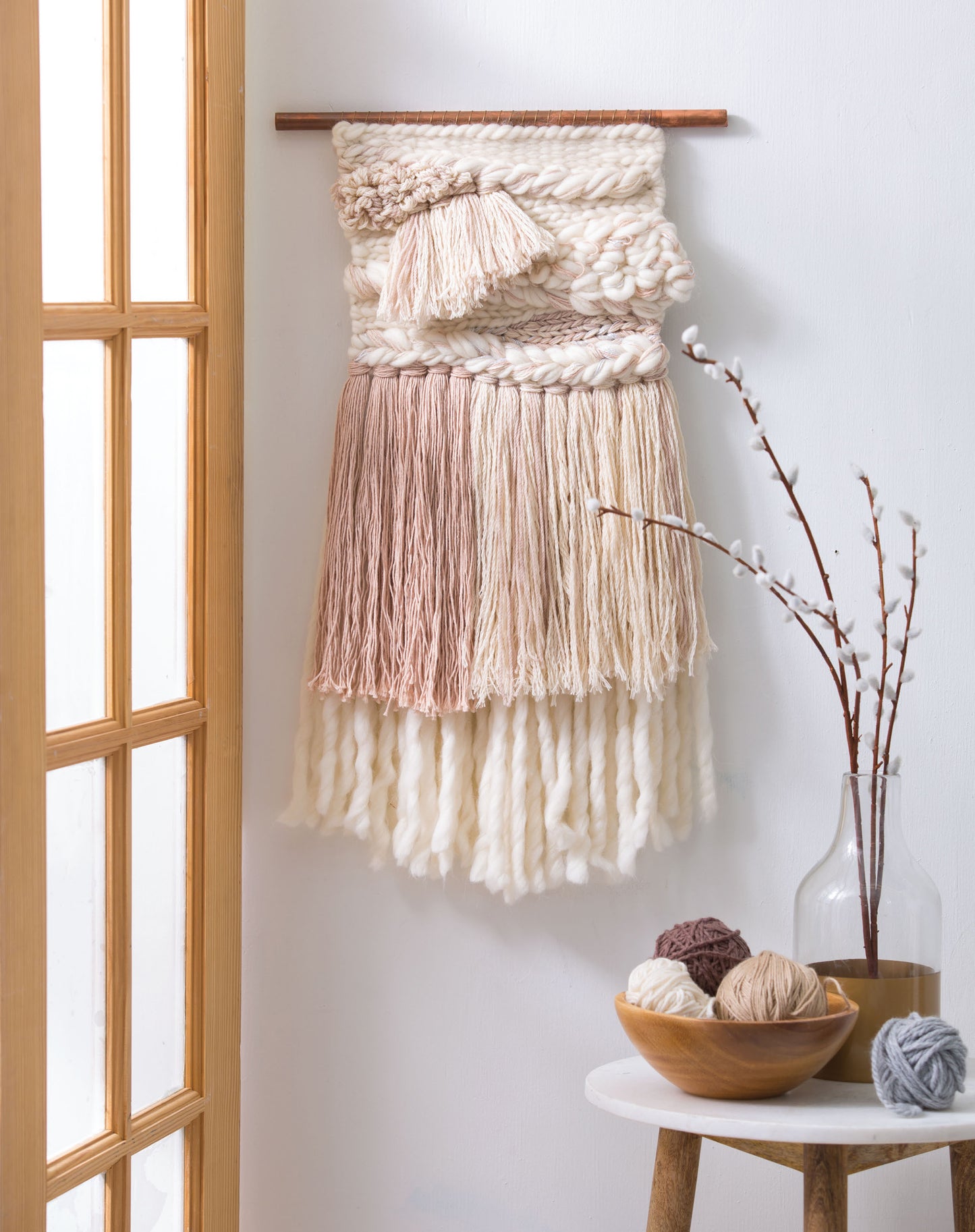 The "Learn to Make Woven Wall Hangings" kit by Leisure Arts, featuring cream and blush yarns, is displayed near a glass door; a small table nearby holds a vase of pussy willow branches and balls of neutral yarn.