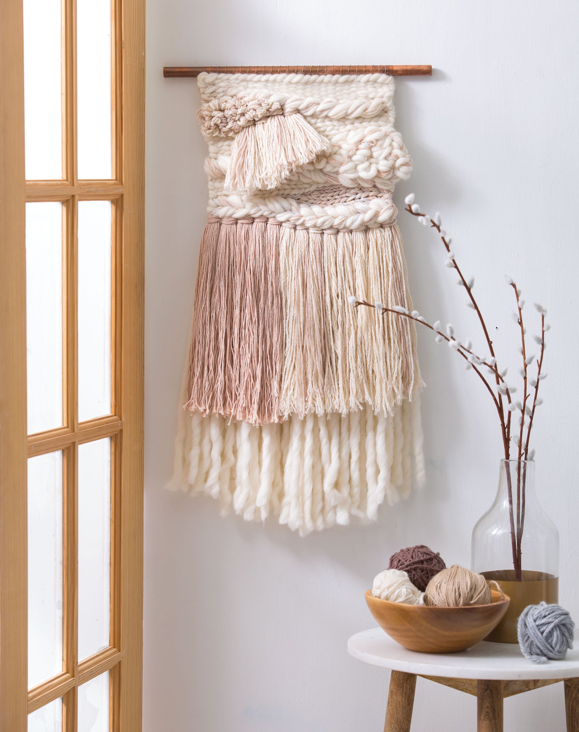 The "Learn to Make Woven Wall Hangings" kit by Leisure Arts, featuring cream and blush yarns, is displayed near a glass door; a small table nearby holds a vase of pussy willow branches and balls of neutral yarn.