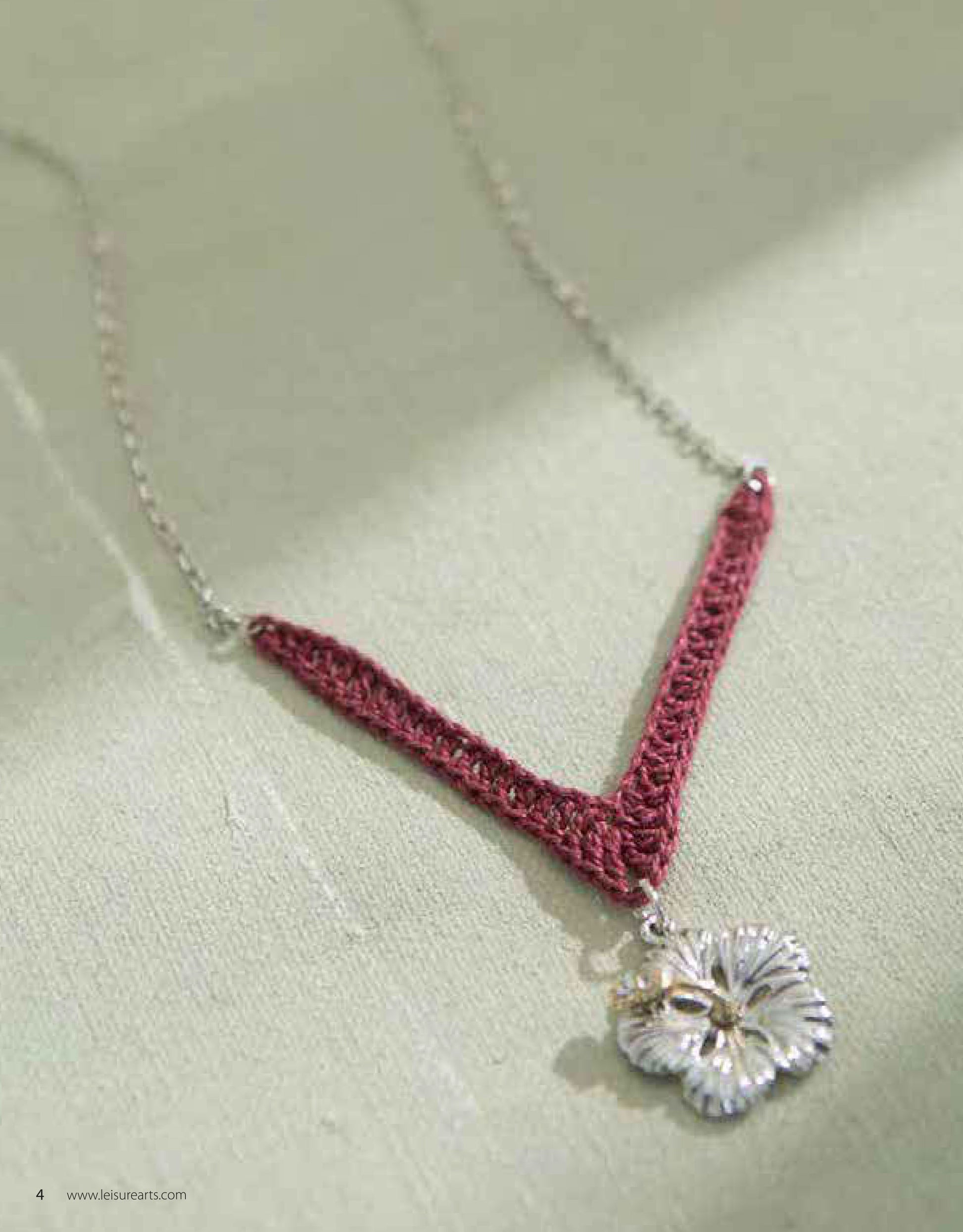 The Jewelry to Crochet necklace by Leisure Arts features a V-shaped burgundy crochet pendant on a silver chain with a silver flower charm, displayed on light green fabric—ideal for crochet jewelry fans.