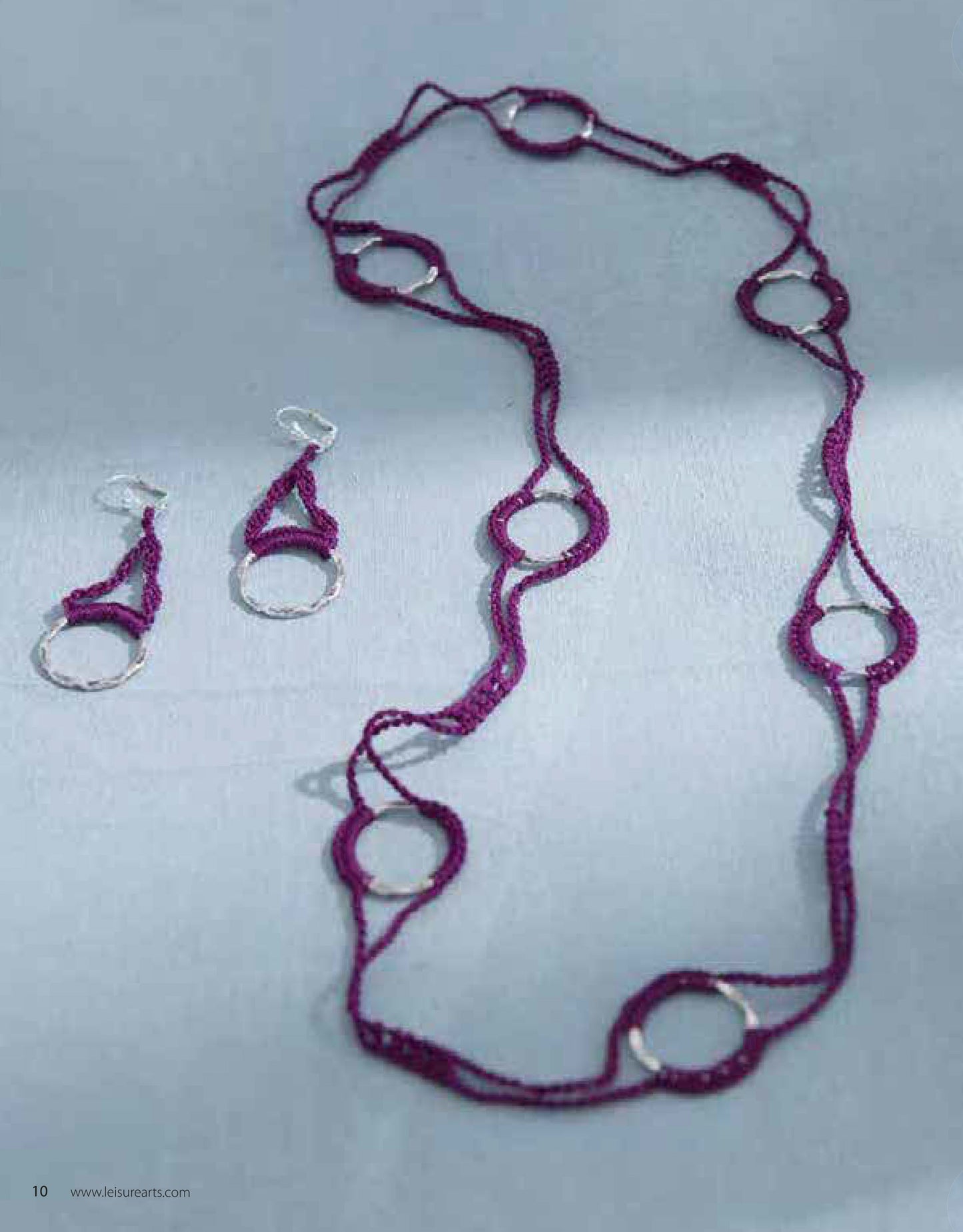 The Jewelry to Crochet set by Leisure Arts features a long purple necklace with silver rings and crochet beads, plus dangling purple earrings with silver hoops, displayed on a light fabric background.