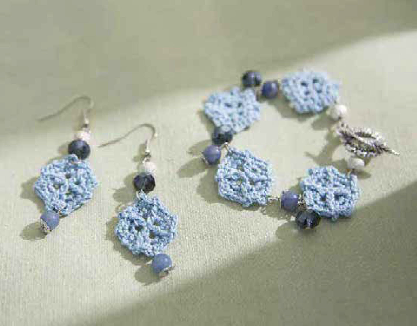 Leisure Arts’ “Jewelry to Crochet” features crocheted blue floral earrings and a matching bracelet with blue beads and a silver leaf charm, elegantly displayed on a light green surface in soft natural light.