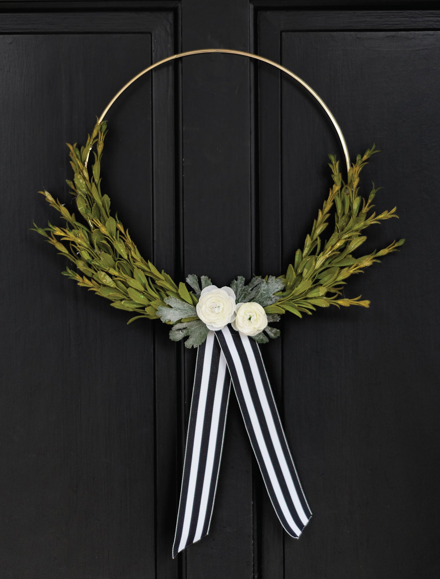 The Hoops Hooray! Digital Download by Leisure Arts features a minimalist wreath with green leaves, two white flowers, and a black-and-white striped ribbon on a thin gold hoop, elegantly hanging on a black door.