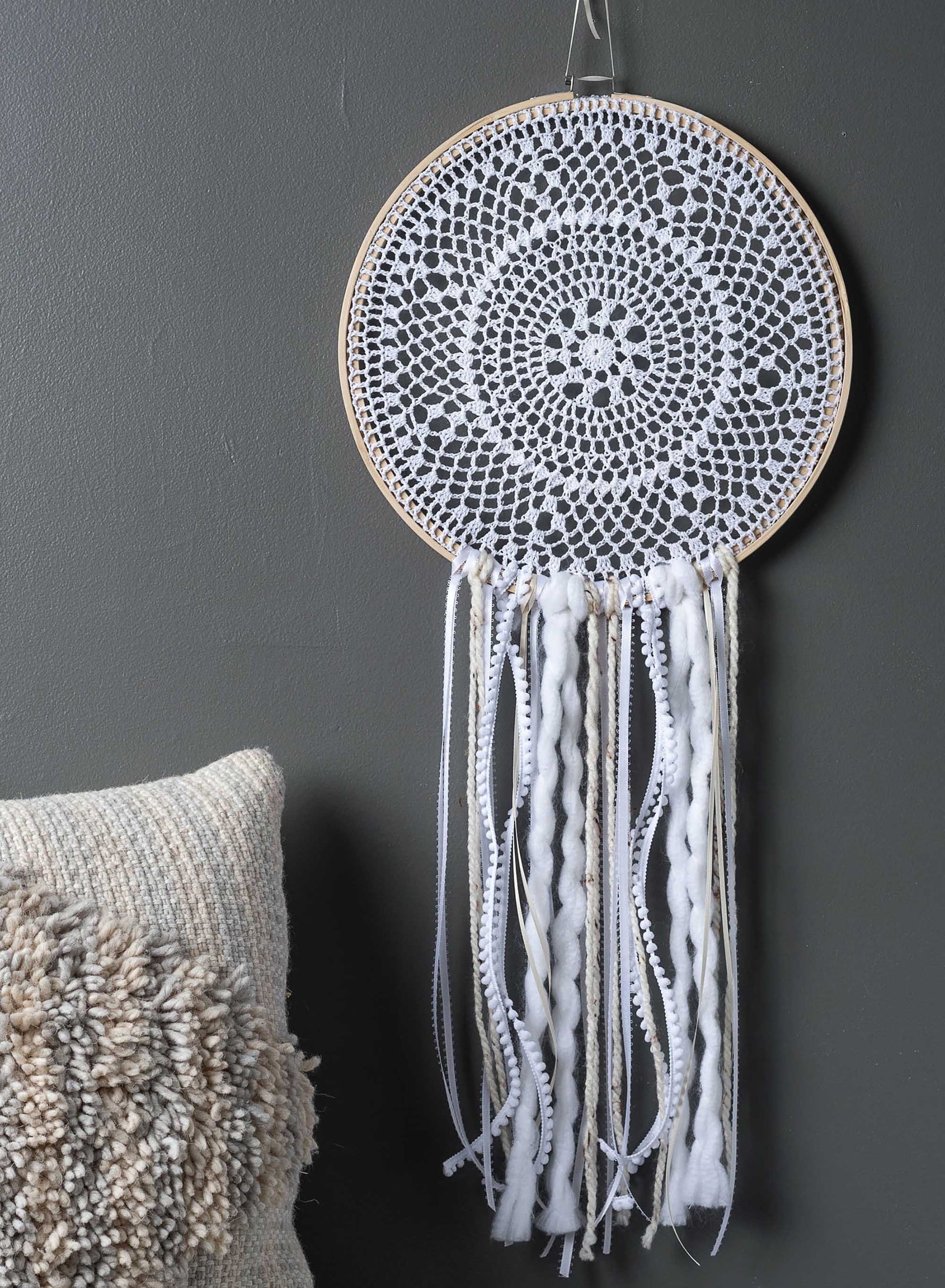 The Hoops Hooray! Digital Download from Leisure Arts features a white crocheted dream catcher with a circular lace design, displayed on a gray wall above a textured beige pillow.