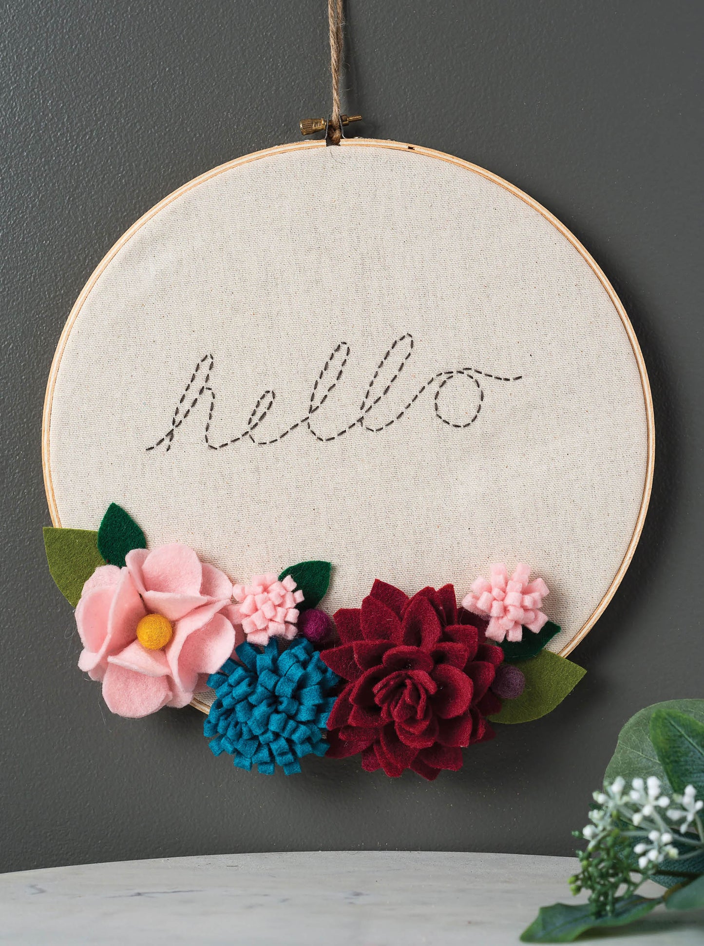 The Hoops Hooray! Digital Download by Leisure Arts features an embroidery hoop on a gray wall with "hello" stitched in black thread, accented by pink, blue, and red felt flowers and green leaves at the bottom for a cheerful display.