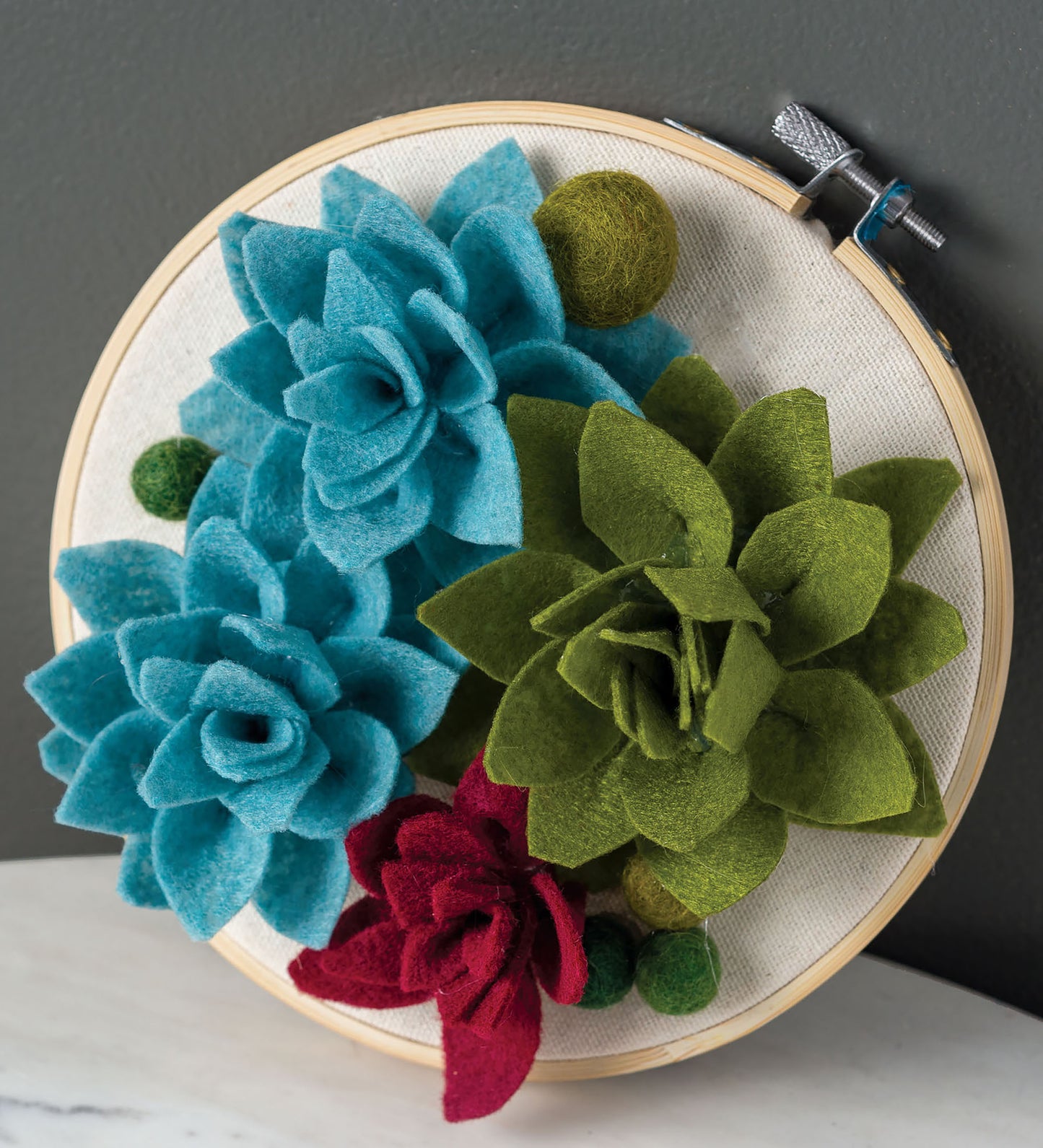 The Hoops Hooray! Digital Download by Leisure Arts features a circular embroidery hoop with handmade felt succulents in blue, green, and red on beige fabric, accented with small green felt balls.