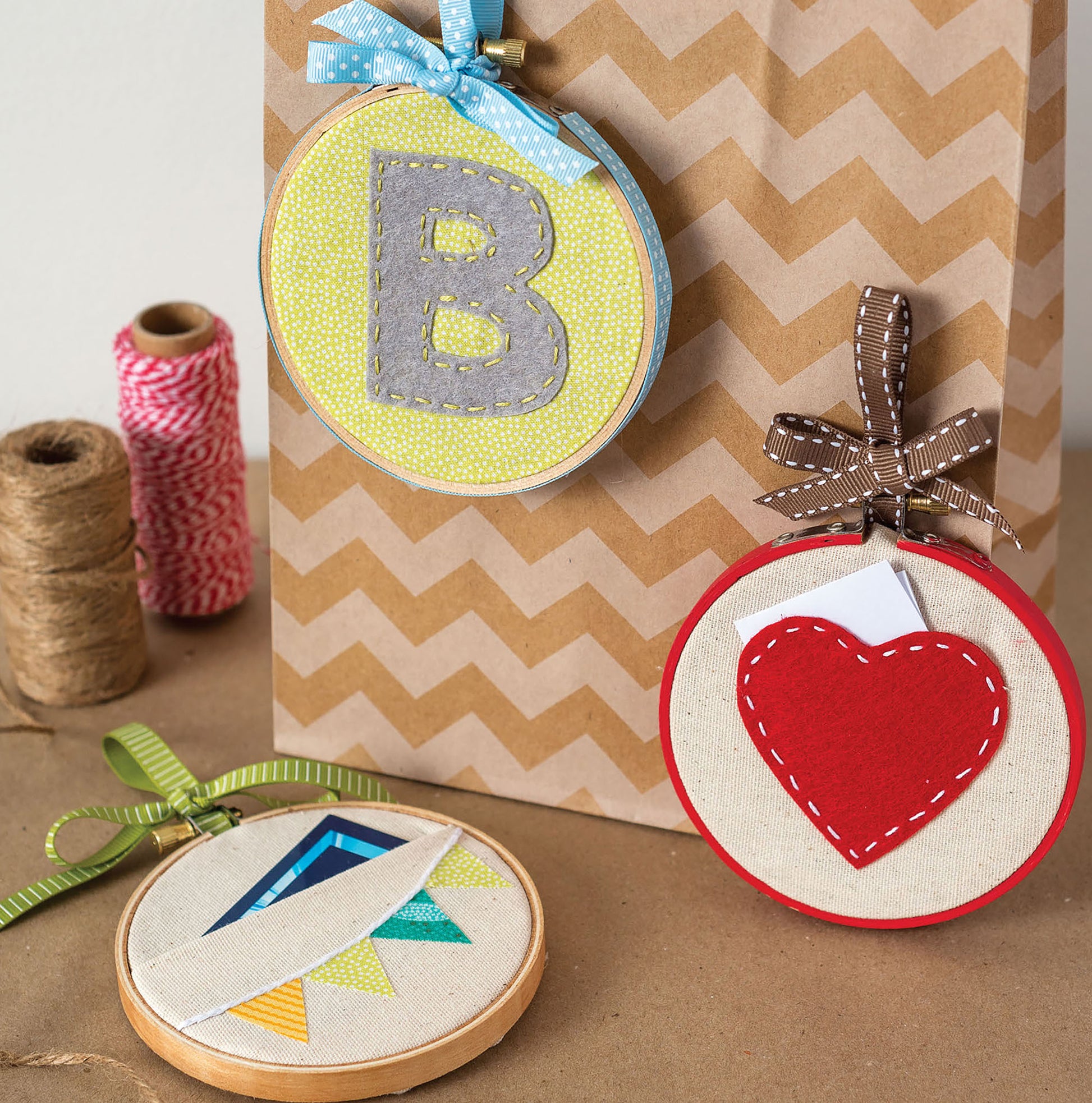 Leisure Arts’ Hoops Hooray! Digital Download features three embroidery hoops—a gray “B,” red heart, and colorful triangles—decorated with bows and displayed on a zigzag gift bag beside twine for a cute handmade touch.