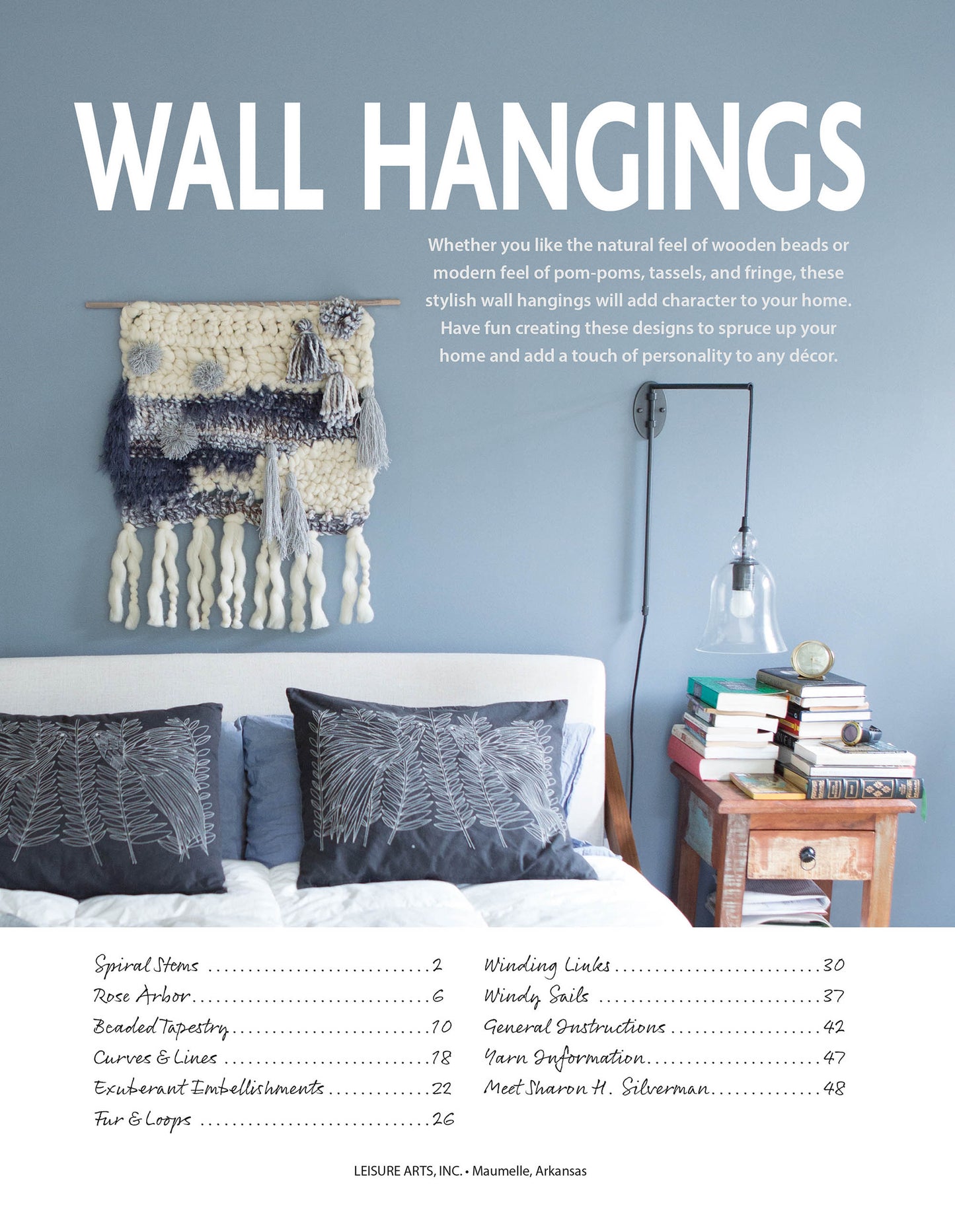 Under the title Wall Hangings—8 Stylish Projects to Define Your Space! by Leisure Arts, a cozy bedroom with a blue accent wall features woven and crochet wall hangings, a hanging light, and a bedside table with books, a cup, and water.