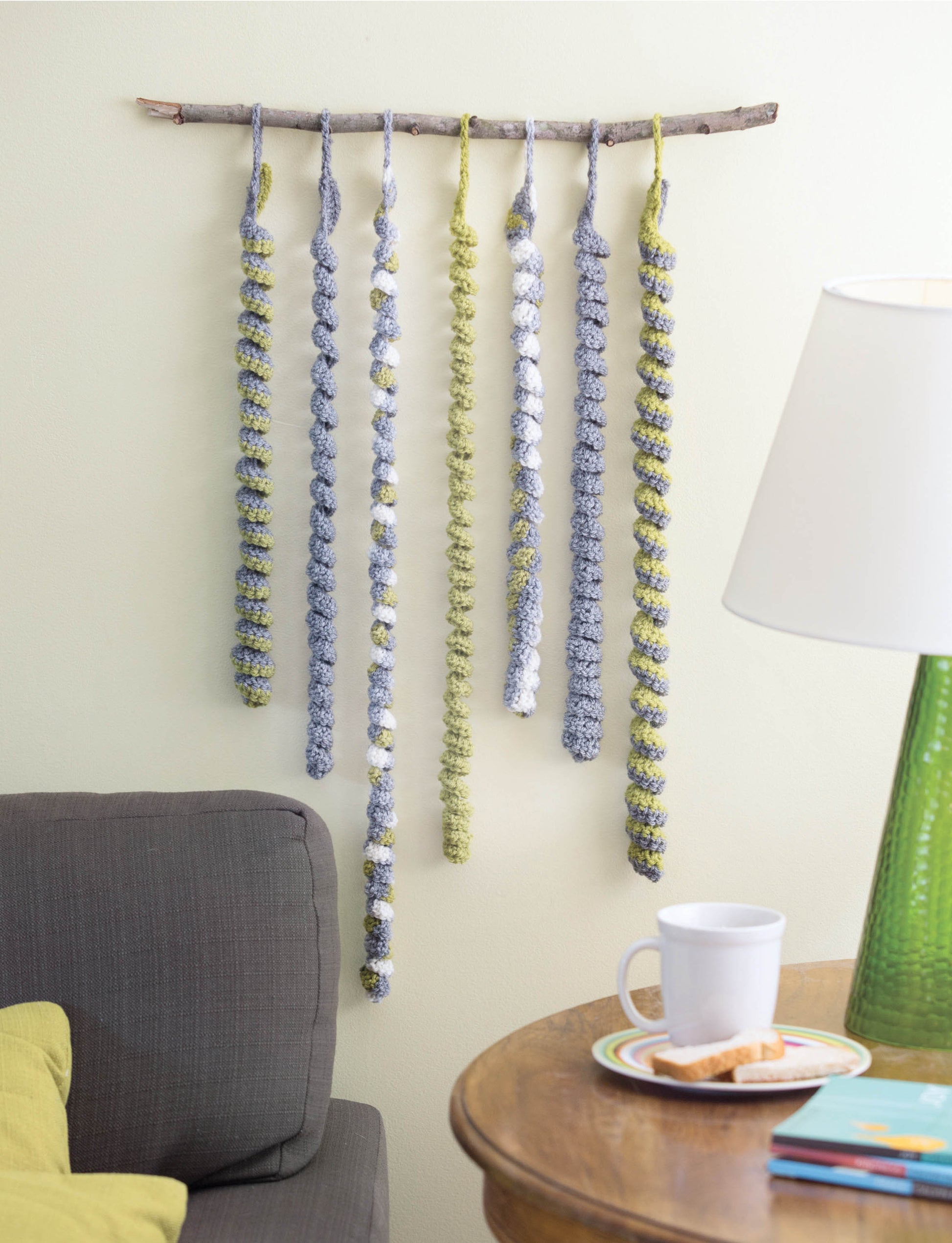A Leisure Arts wall hanging from "Wall Hangings—8 Stylish Projects to Define Your Space!" features six long, curly yarn strands in gray, green, and white. Beneath it sits a gray sofa with green pillows and a round table set with a lamp and snacks.