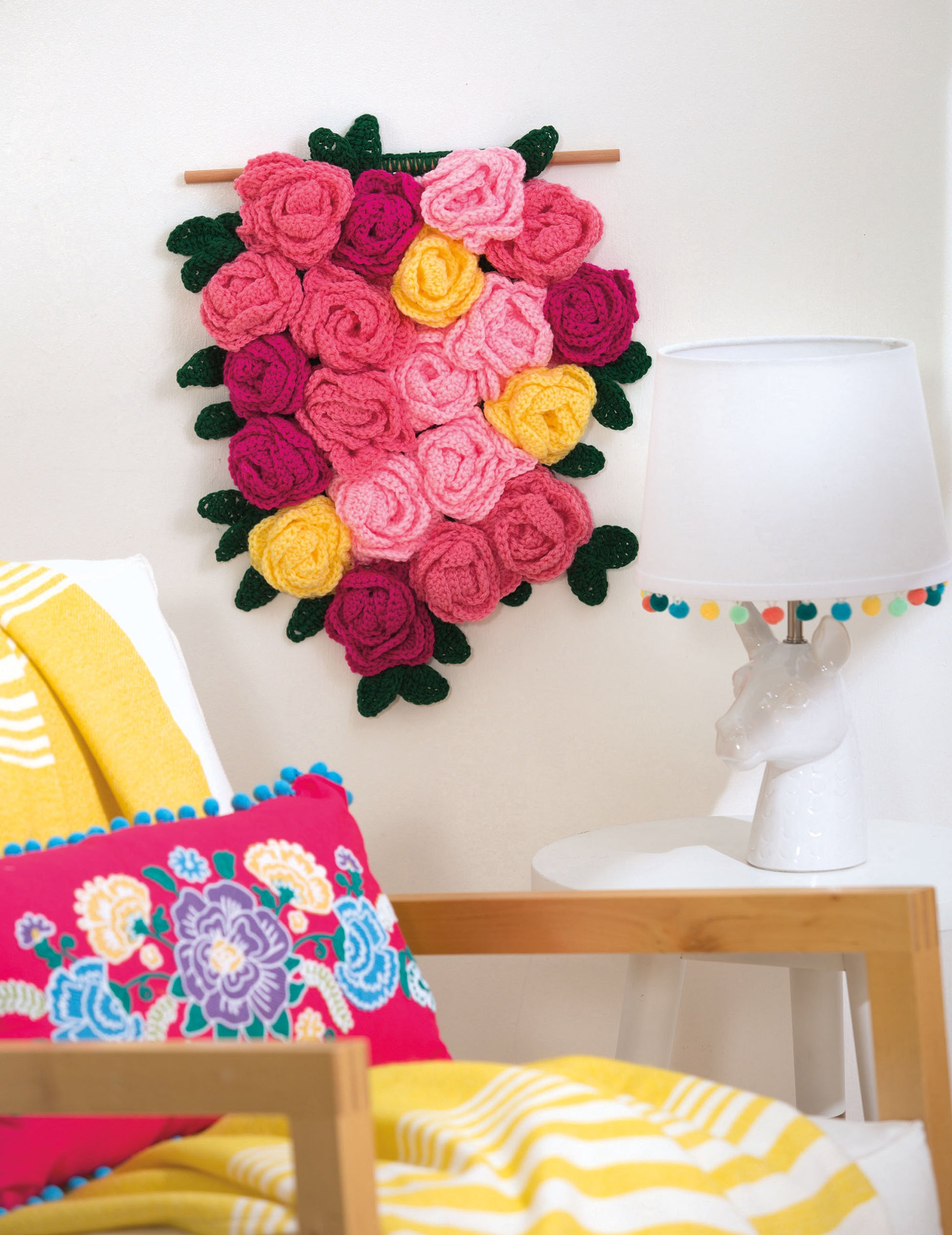 A cozy bedroom corner features a white giraffe-shaped lamp, a colorful embroidered pillow, a yellow blanket, and Leisure Arts' "Wall Hangings—8 Stylish Projects to Define Your Space! Digital Download" with crocheted roses and green leaves.
