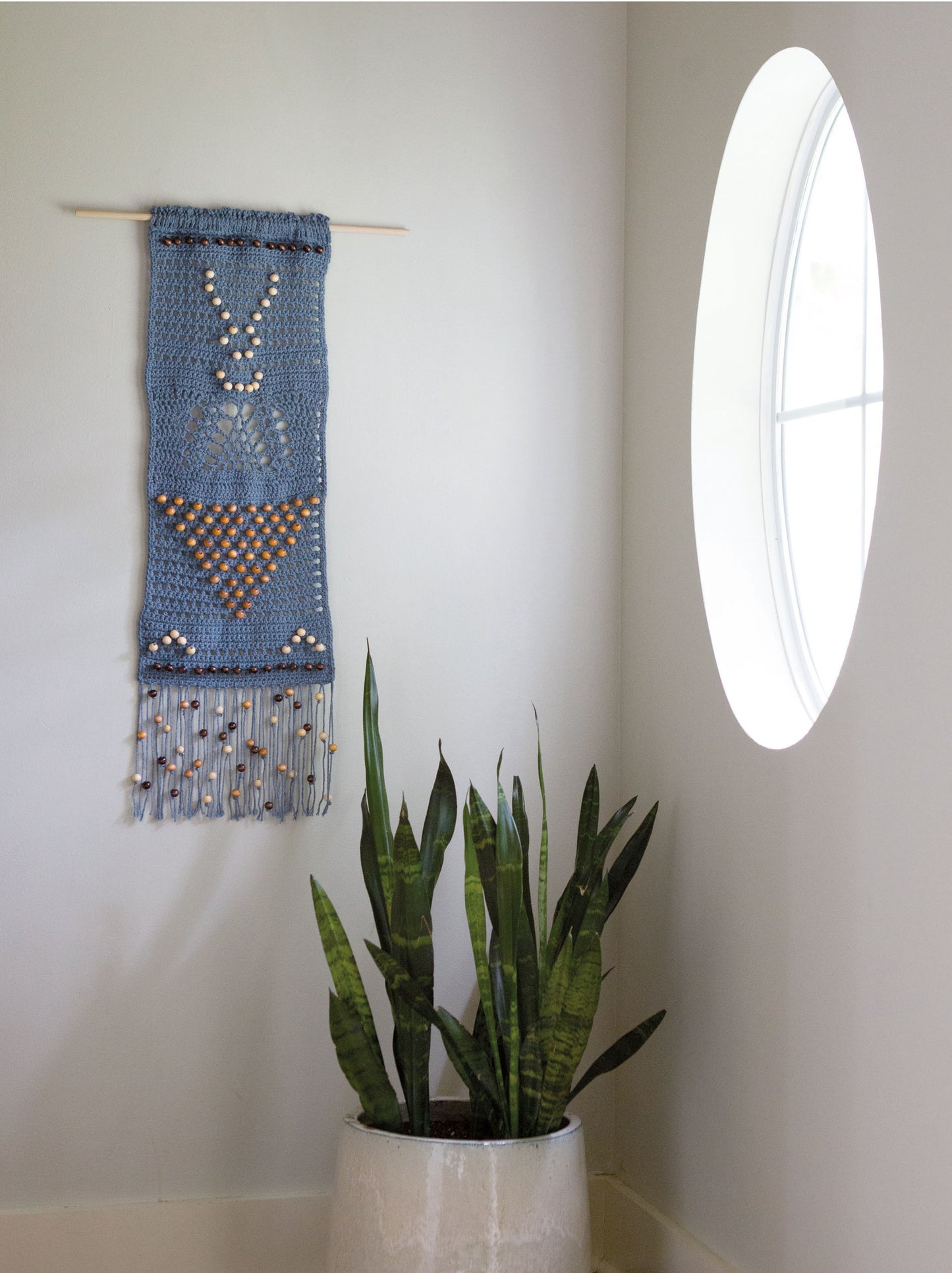 In a sunlit room, a potted snake plant rests near an oval window, while the white wall above features a beaded, fringed crochet piece made with Leisure Arts' Wall Hangings—8 Stylish Projects to Define Your Space! digital download.
