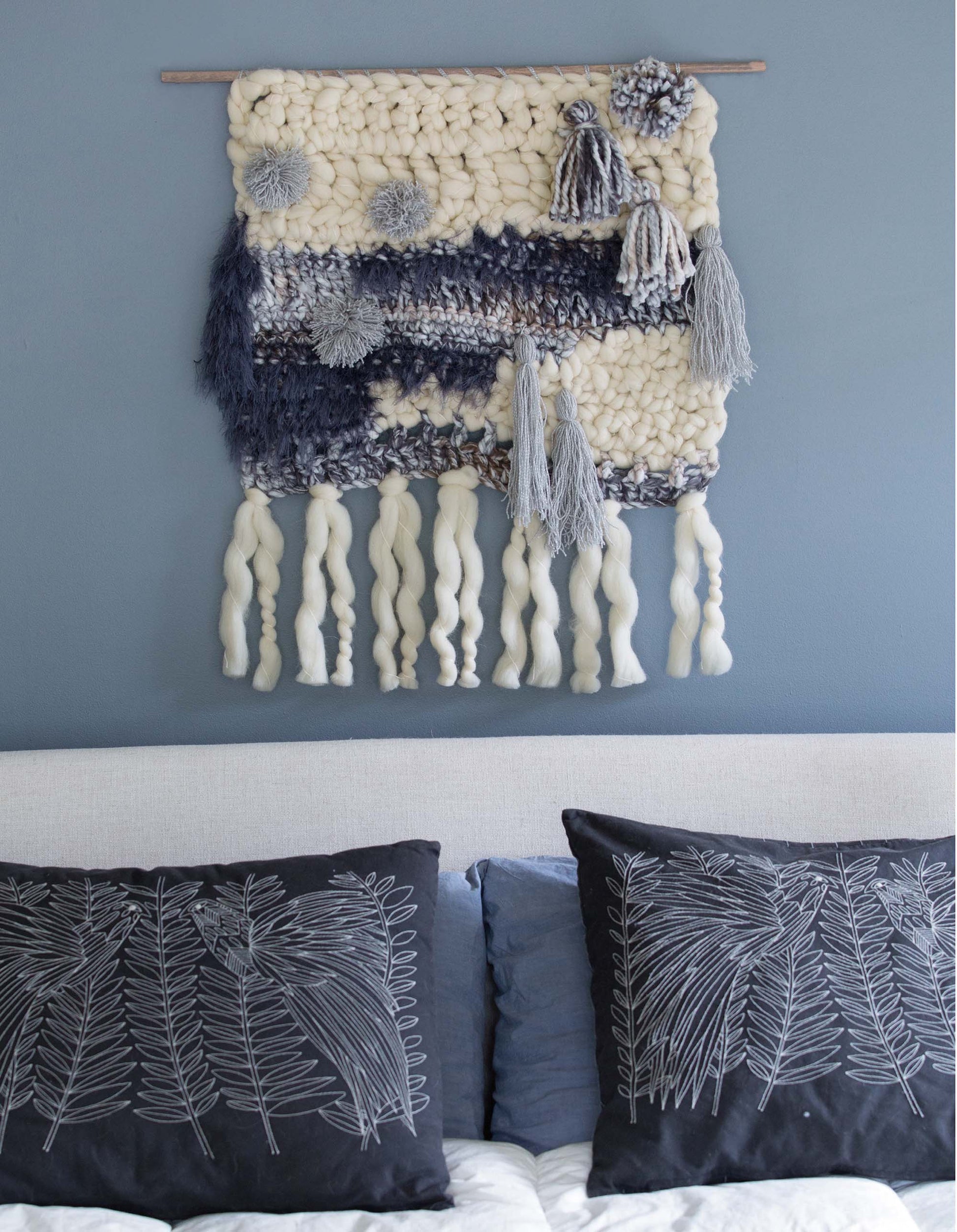 Elevate your space with "Wall Hangings—8 Stylish Projects to Define Your Space!" by Leisure Arts, a digital download featuring cream and gray yarn designs with tassels and pom-poms, perfect above a bed with embroidered botanical pillows.