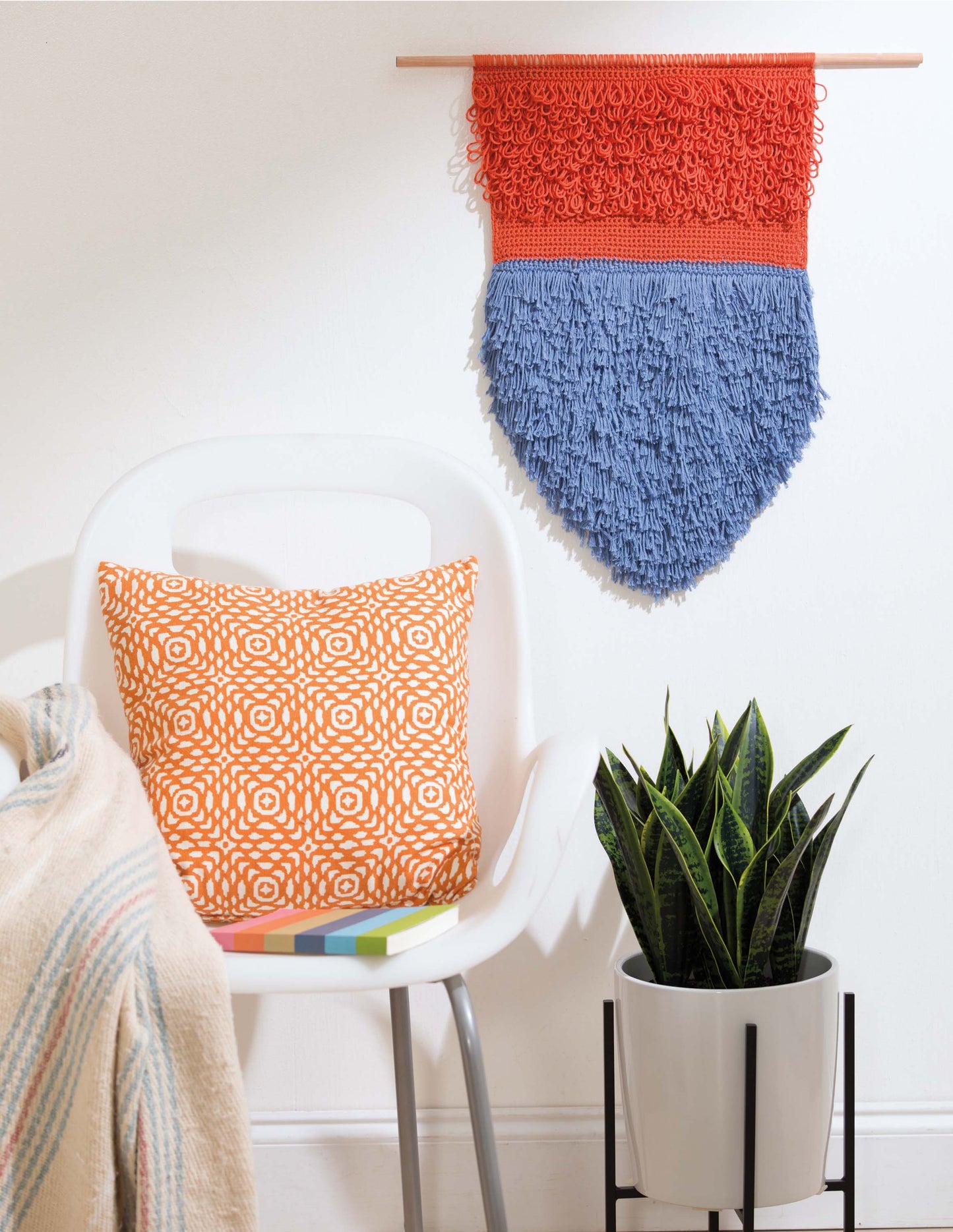A modern white chair with an orange pillow sits by a potted snake plant; above, a vibrant crochet from Leisure Arts’ “Wall Hangings—8 Stylish Projects to Define Your Space! Digital Download” adds flair to the white wall.
