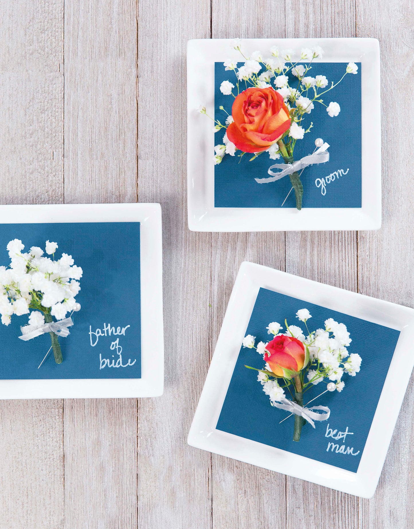 Three white square plates on a light wood surface display blue napkins reading “father of bride,” “groom,” and “best man” in white script, topped with boutonnieres—perfect personalized Handmade Wedding decor by Leisure Arts.