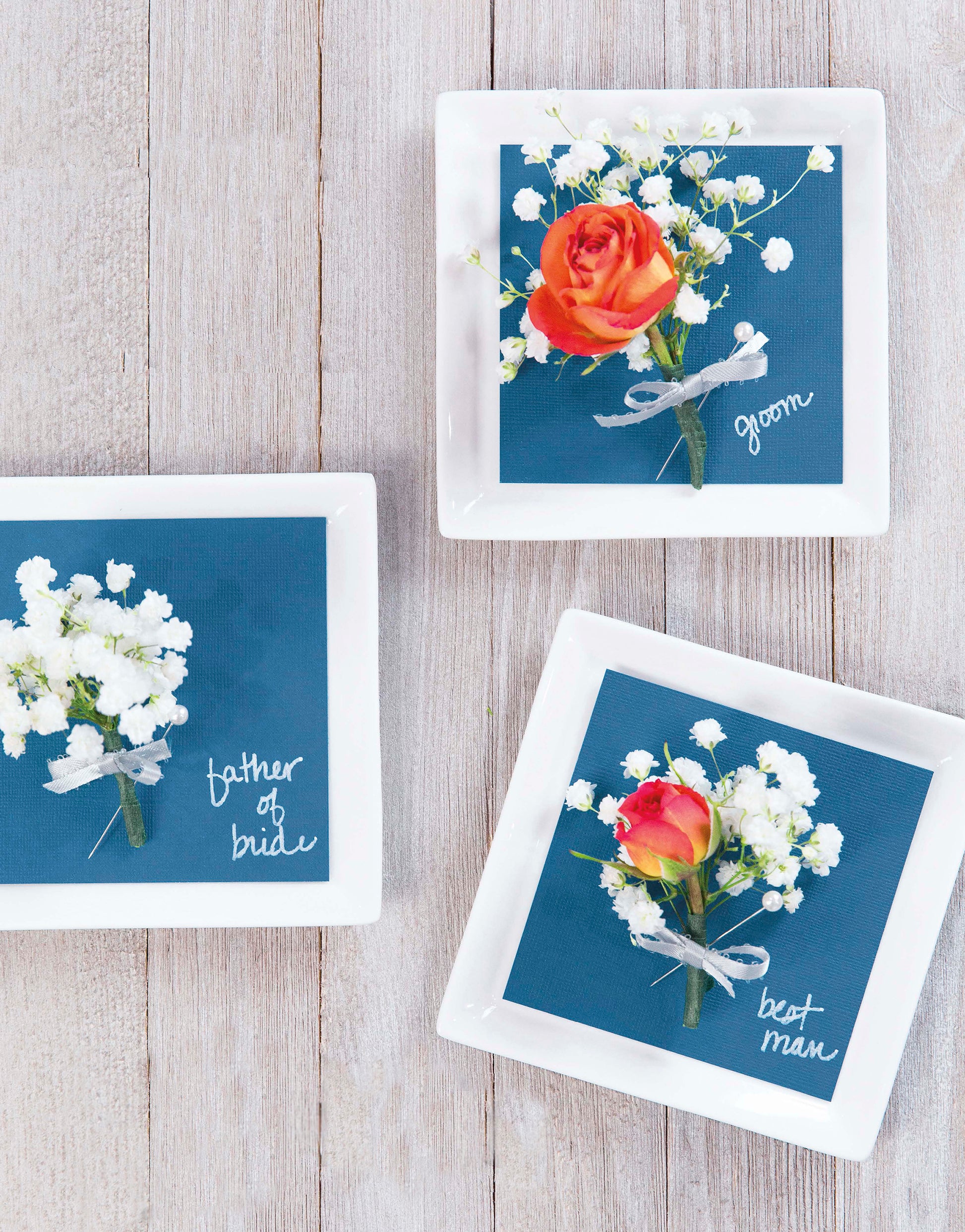 Three white square plates on a light wood surface display blue napkins reading “father of bride,” “groom,” and “best man” in white script, topped with boutonnieres—perfect personalized Handmade Wedding decor by Leisure Arts.