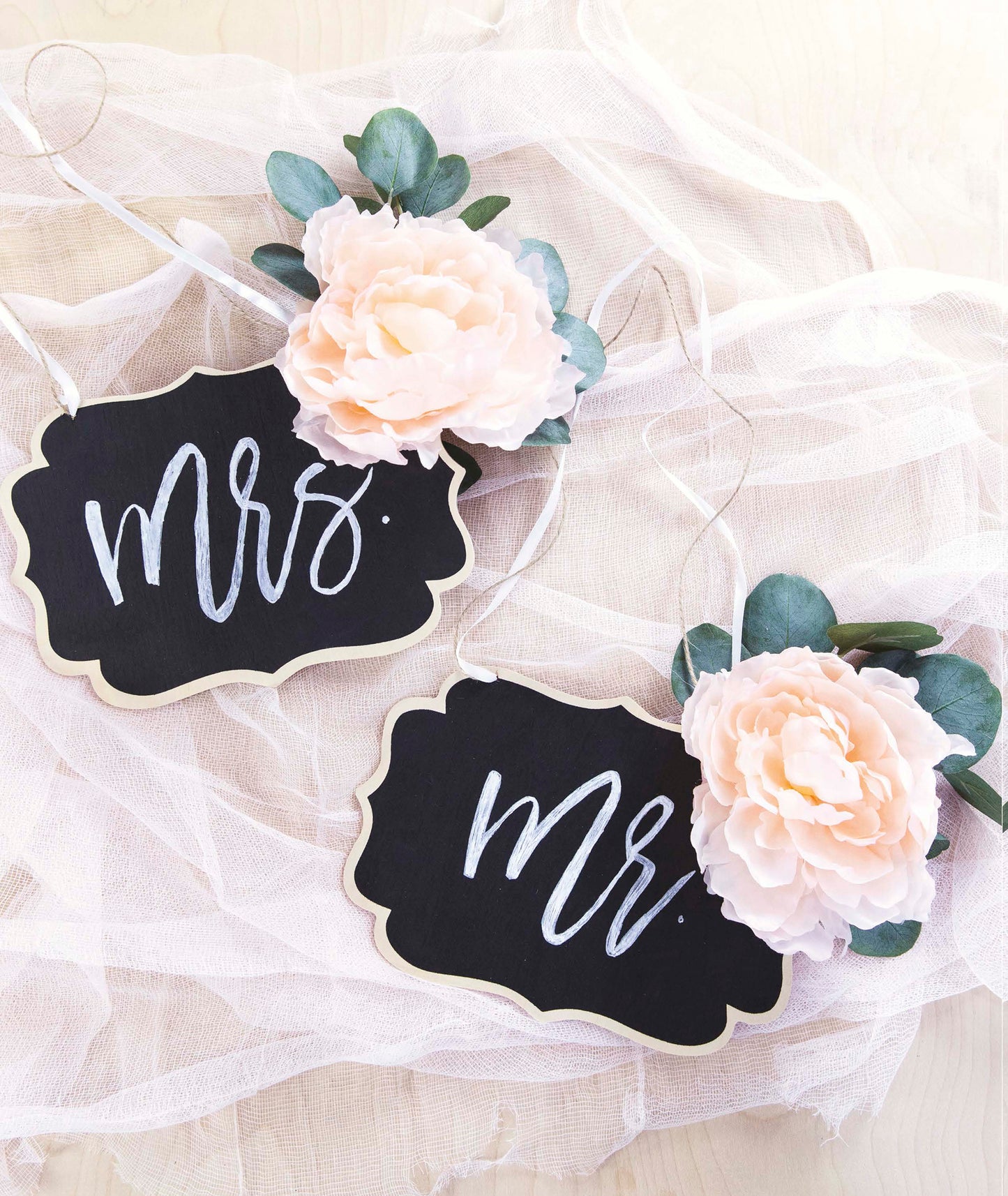 The Handmade Wedding by Leisure Arts features two black chalkboard chair signs with "Mrs." and "Mr." in white calligraphy, each adorned with a blush peony and eucalyptus—perfect personalized wedding decor displayed on white tulle.