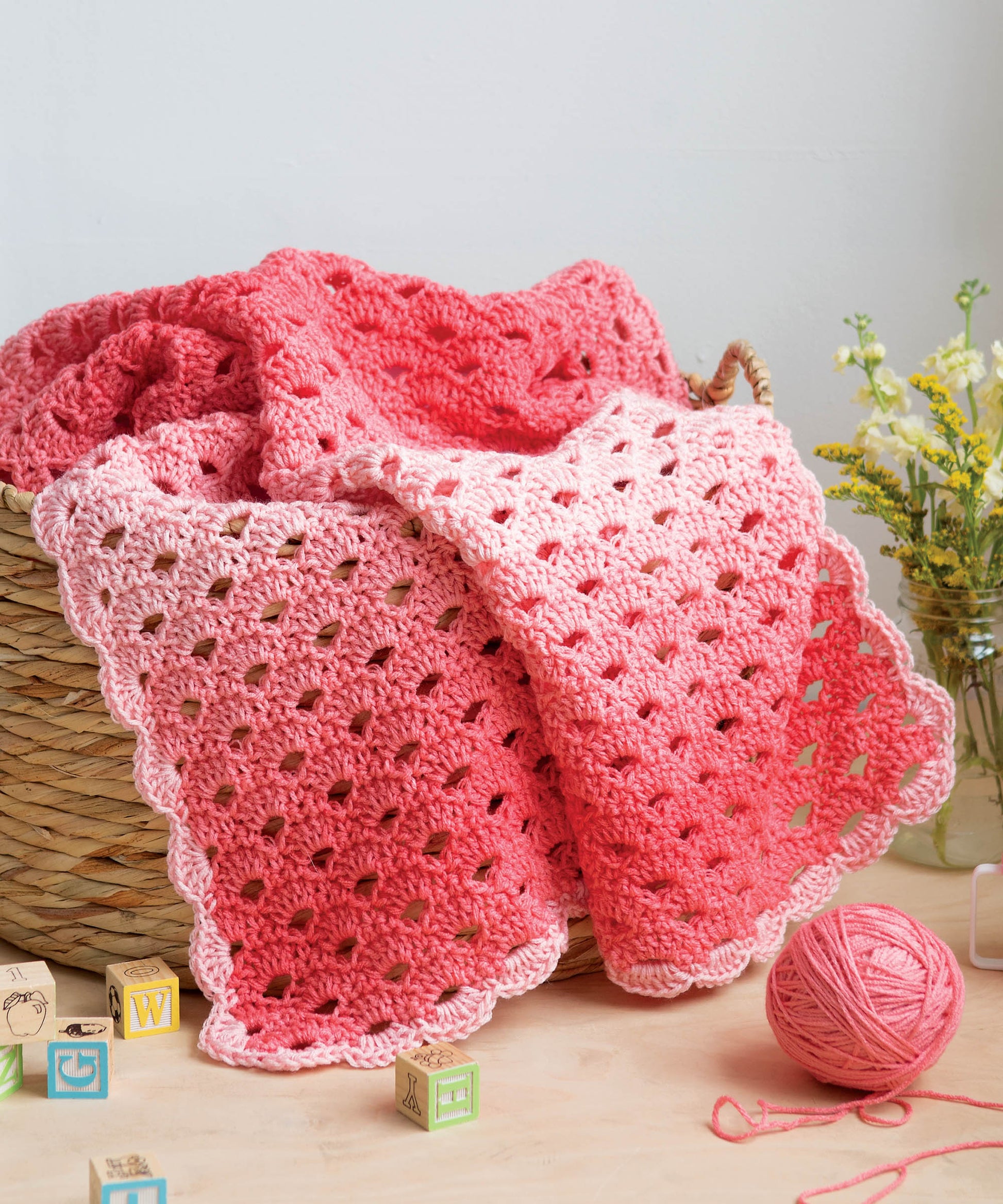 A crochet blanket made with Leisure Arts' Crochet Self-Striping Projects yarn in pink and coral rests on a basket, with a matching yarn ball and alphabet blocks nearby. Yellow flowers add charm, making it perfect for cozy creations.