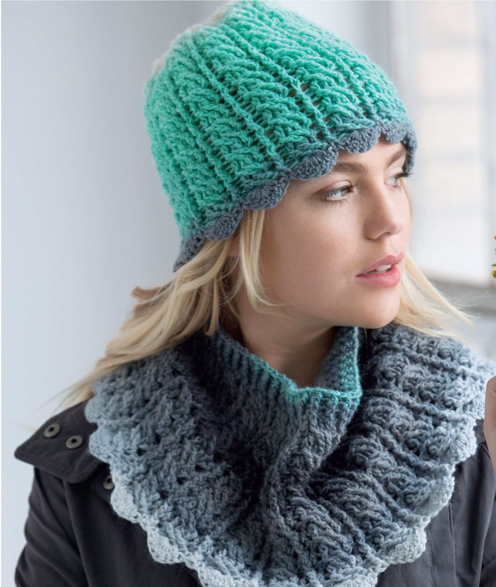 A woman wearing a teal knit hat with a gray scalloped edge and matching scarf, both made with ombre yarn, models patterns from Leisure Arts’ Crochet Self-Striping Projects as she looks to the side indoors.
