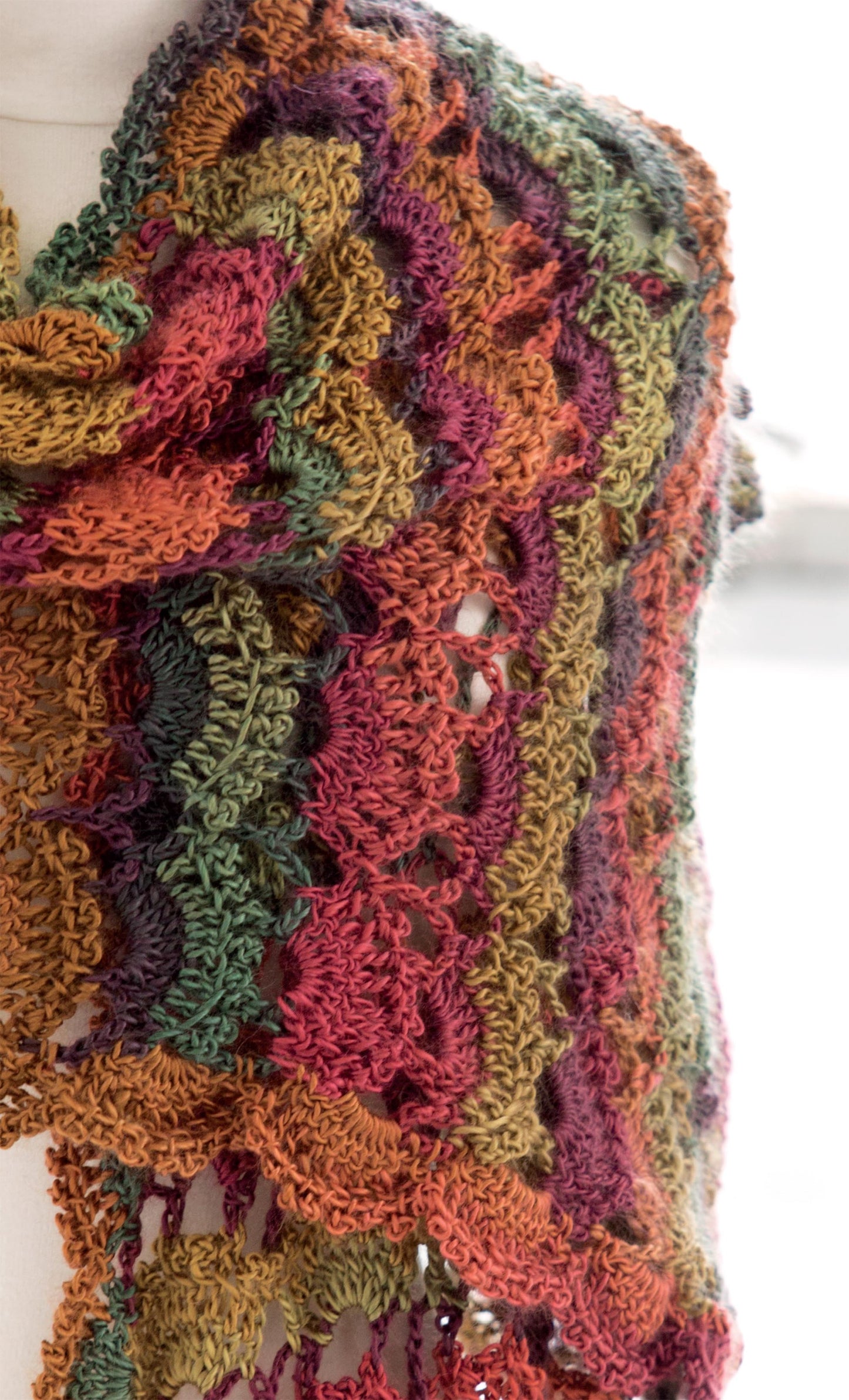 A close-up of a wavy openwork shawl from Leisure Arts’ Crochet Self-Striping Projects, showing vibrant green, orange, red, and purple stripes, draped over a light surface.