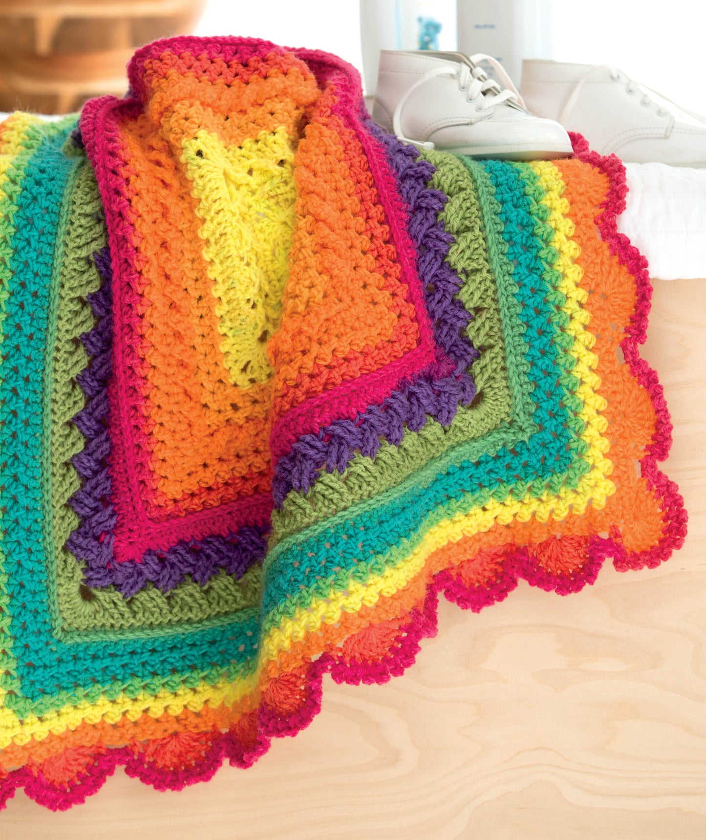 A vibrant crocheted blanket from Leisure Arts' Crochet Self-Striping Projects displays rows of yellow, orange, red, pink, green, and purple yarn, draped on light wood with white baby shoes in the background.