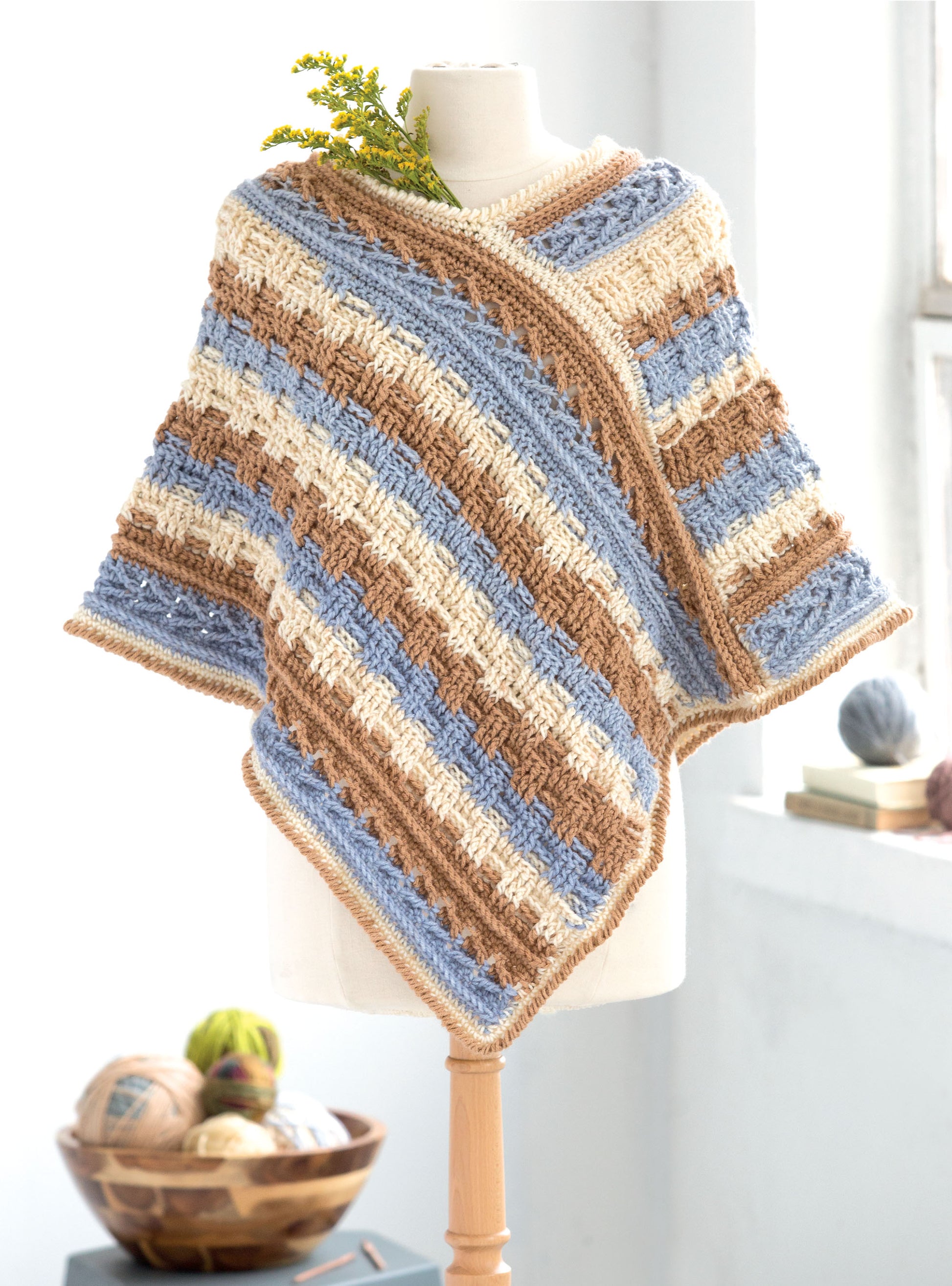 A mannequin models a poncho made from blue, cream, and brown self-striping yarn from Leisure Arts’ Crochet Self-Striping Projects. Yellow flowers accent the neckline; yarn balls rest in a bowl on the windowsill.