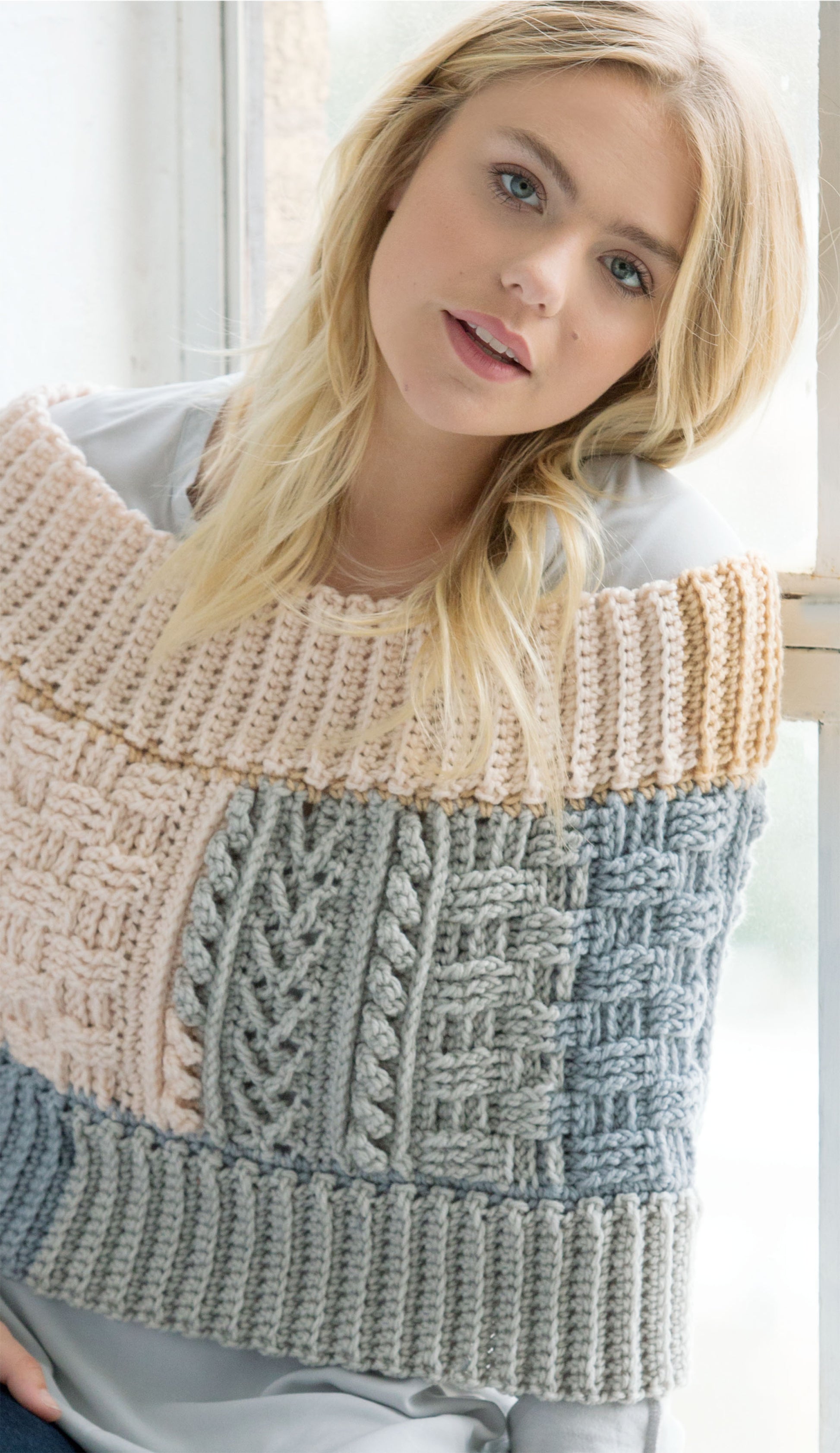 A young woman with blonde hair sits by a window, wearing a multicolored sweater made from self-striping yarn, featured in Crochet Self-Striping Projects by Leisure Arts. She gazes softly at the camera with a gentle smile.