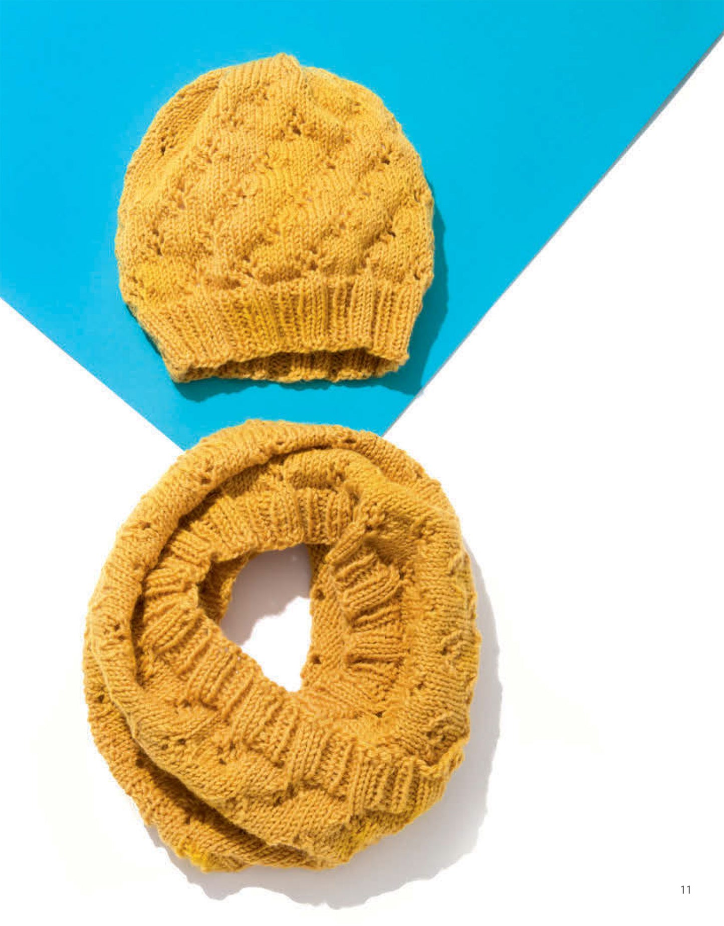 Leisure Arts Knit Accessories, featuring a yellow knitted hat and matching infinity scarf, are shown on a white surface with a blue triangle backdrop—perfect for adding cozy style to your look.