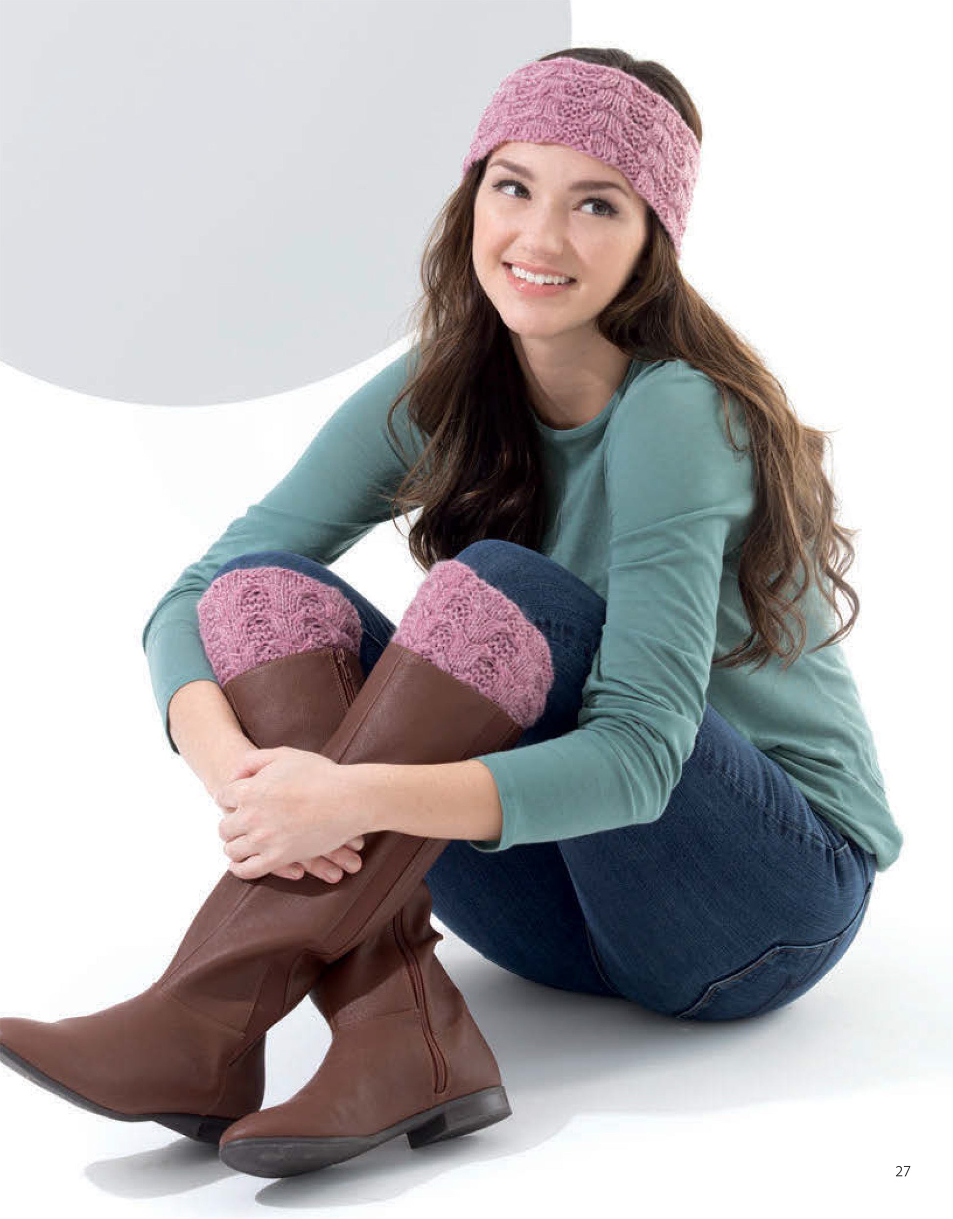 A woman wearing a light teal long-sleeve shirt and blue jeans smiles as she shows off her matching pink Knit Accessories—headband and boot cuffs with cozy winter patterns—from Leisure Arts.