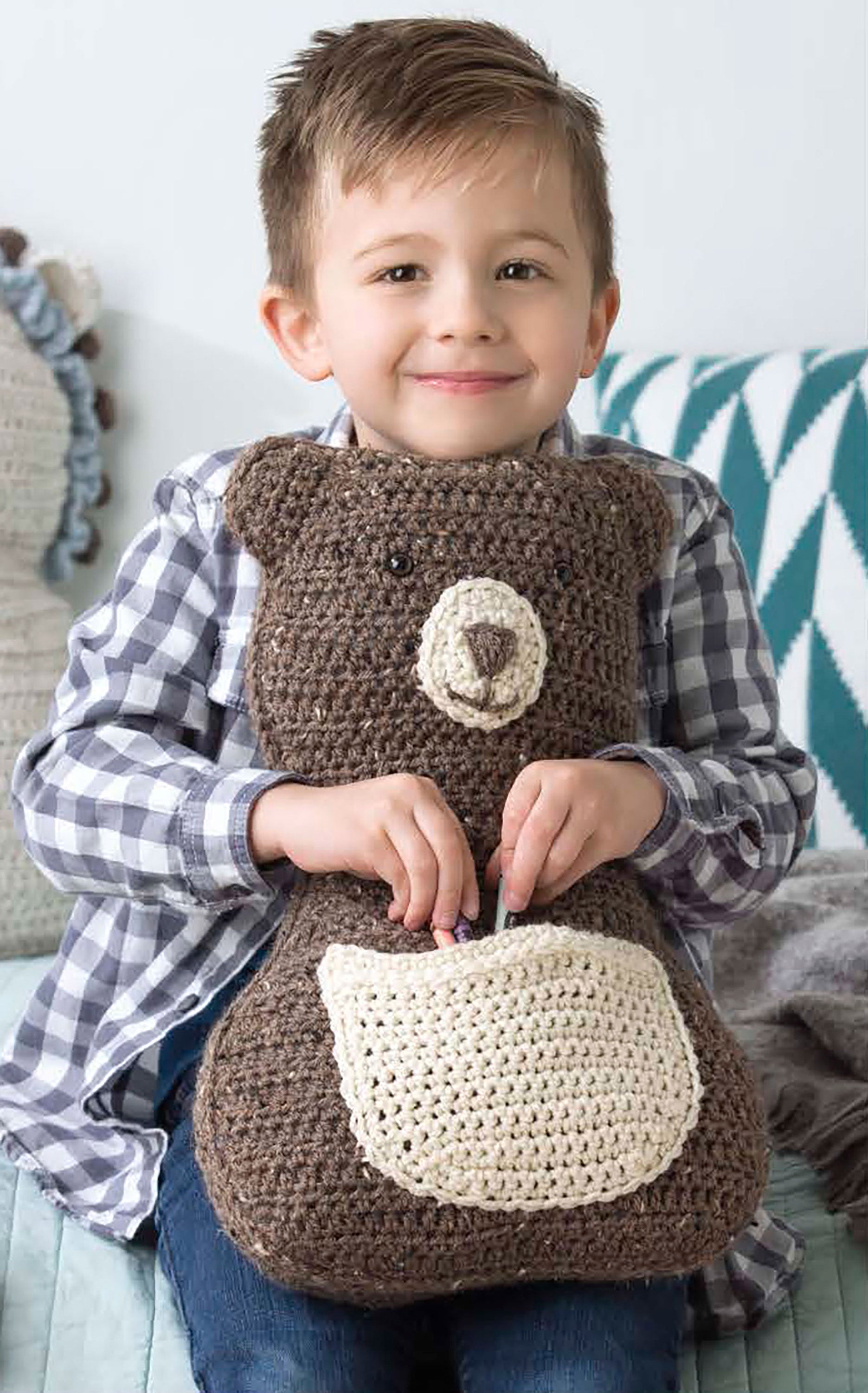 A smiling young boy with short brown hair and a plaid shirt hugs a large crocheted brown bear pillow—one of the Fun Animal Pillows from Leisure Arts’ “9 Huggable Friends to Stitch for Little Ones”—while sitting on the couch.