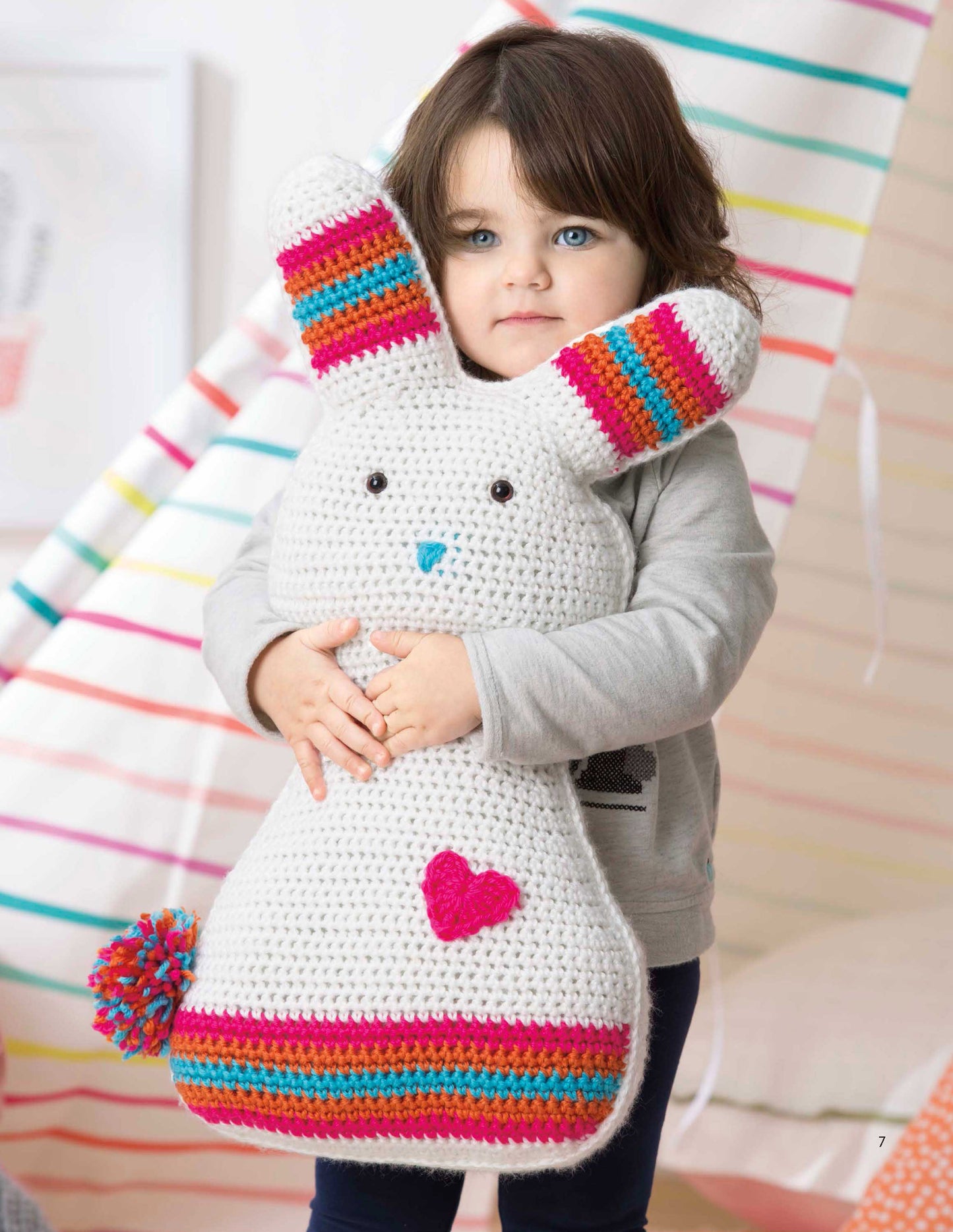 A young child with blue eyes and brown hair hugs a large crocheted white bunny, one of our Fun Animal Pillows from Leisure Arts' "9 Huggable Friends to Stitch for Little Ones," featuring colorful stripes and a heart on its body.