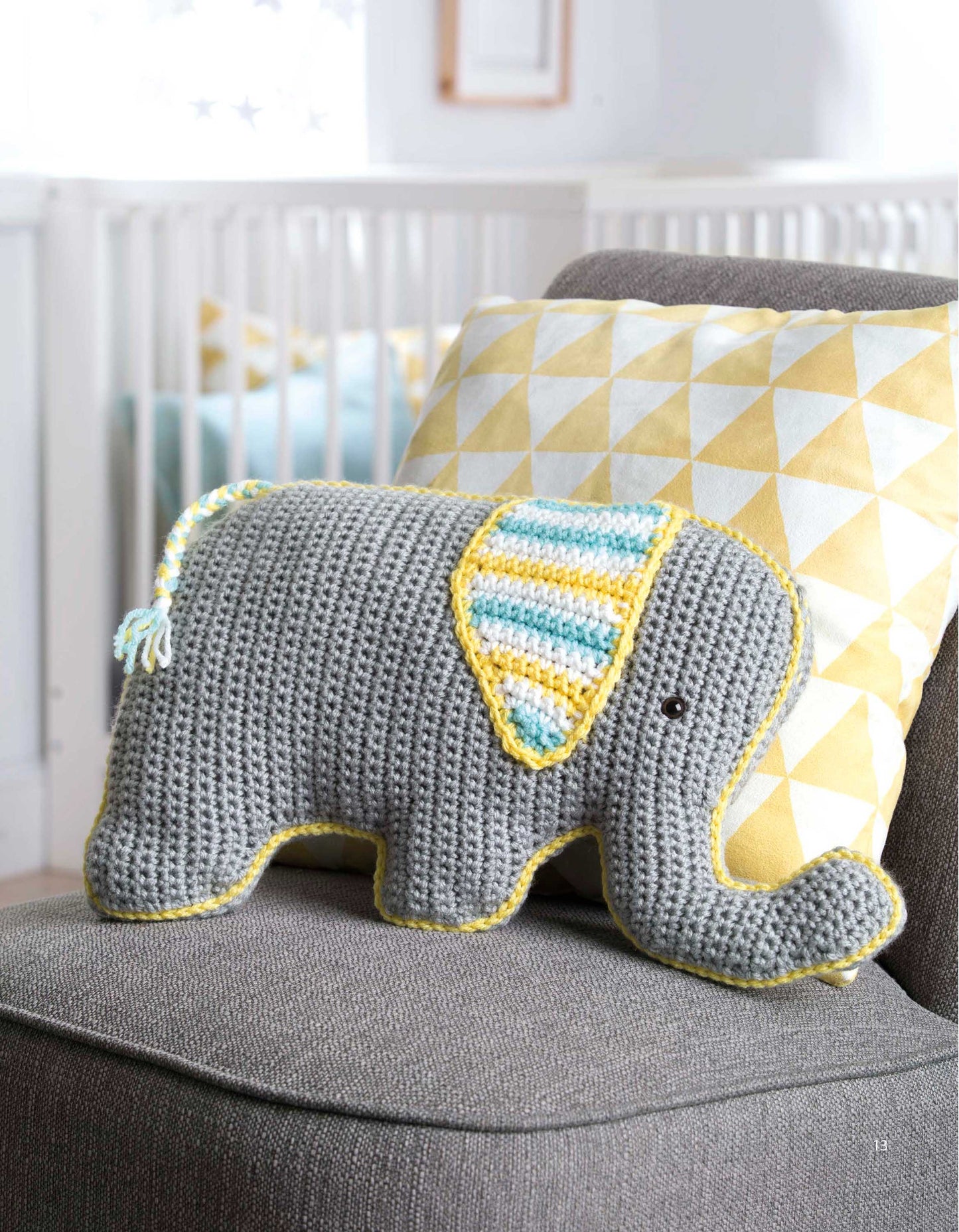 A gray crocheted elephant pillow from Leisure Arts' Fun Animal Pillows—9 Huggable Friends to Stitch for Little Ones, with yellow and blue striped ears and tail, sits on a gray chair by a yellow and white geometric cushion.
