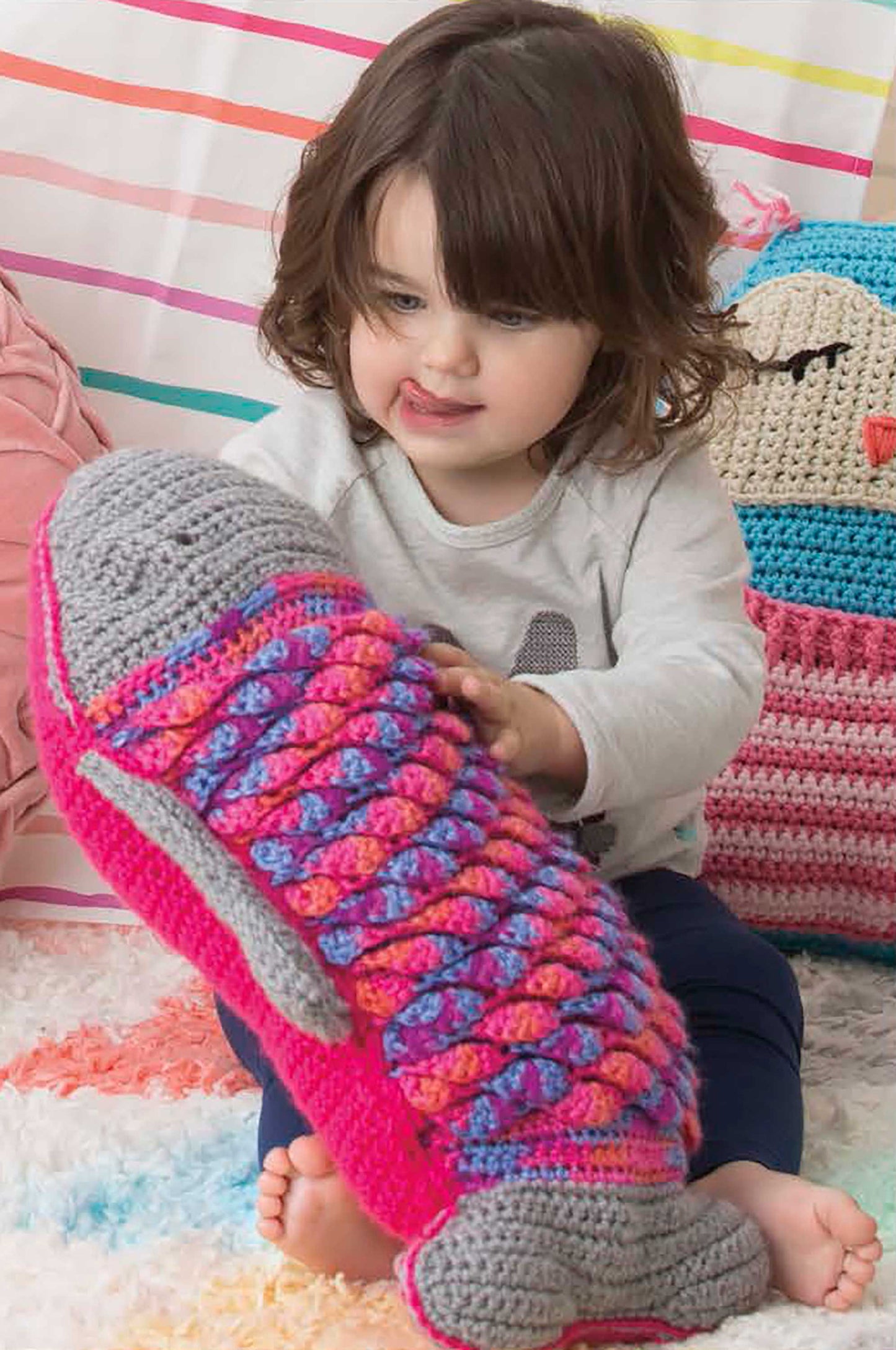 A young child sits on a colorful rug, hugging a fish-shaped pillow from Leisure Arts' Fun Animal Pillows—9 Huggable Friends to Stitch for Little Ones, surrounded by other cute animal pillows and a striped background for extra coziness.