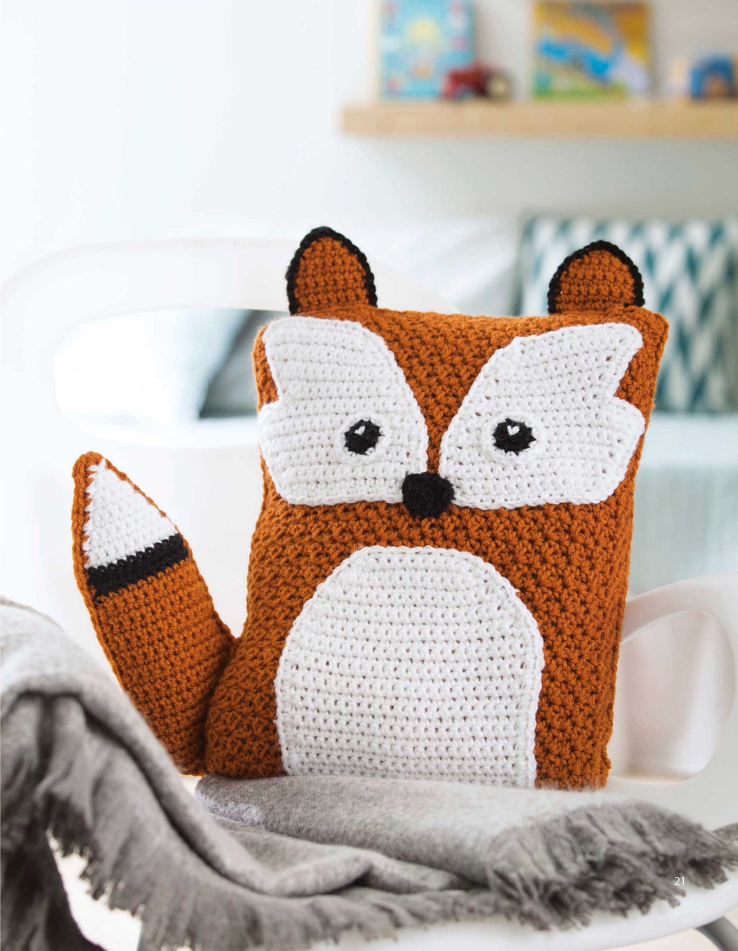 Add charm to any space with Leisure Arts’ Fun Animal Pillows—9 Huggable Friends to Stitch for Little Ones. This crocheted fox cushion brightens up a cozy room, sitting on a white chair beside a gray blanket.