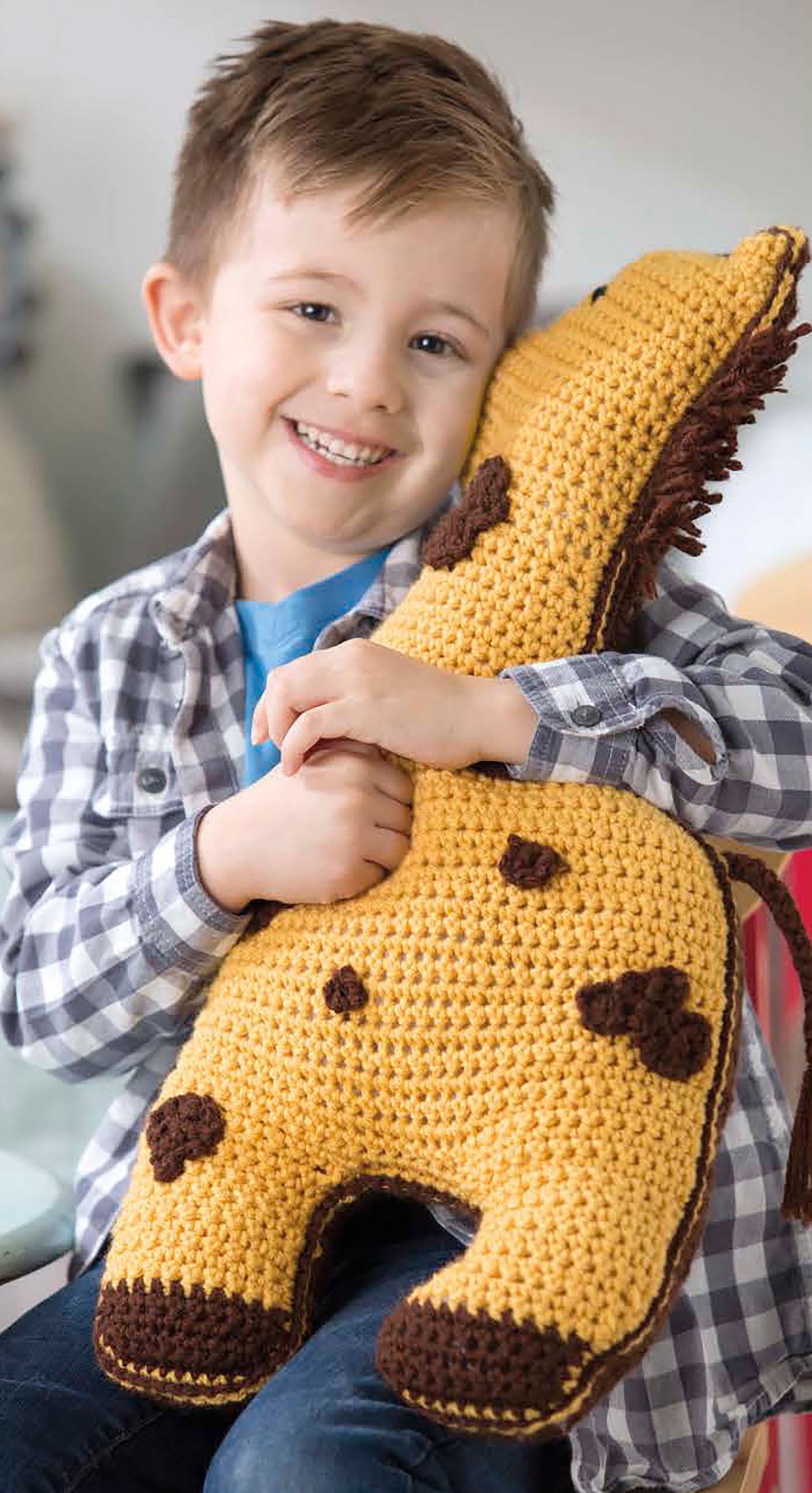 A smiling young boy in a plaid shirt hugs a yellow giraffe plush from Leisure Arts’ “Fun Animal Pillows—9 Huggable Friends to Stitch for Little Ones,” one of his favorite crochet animal buddies.