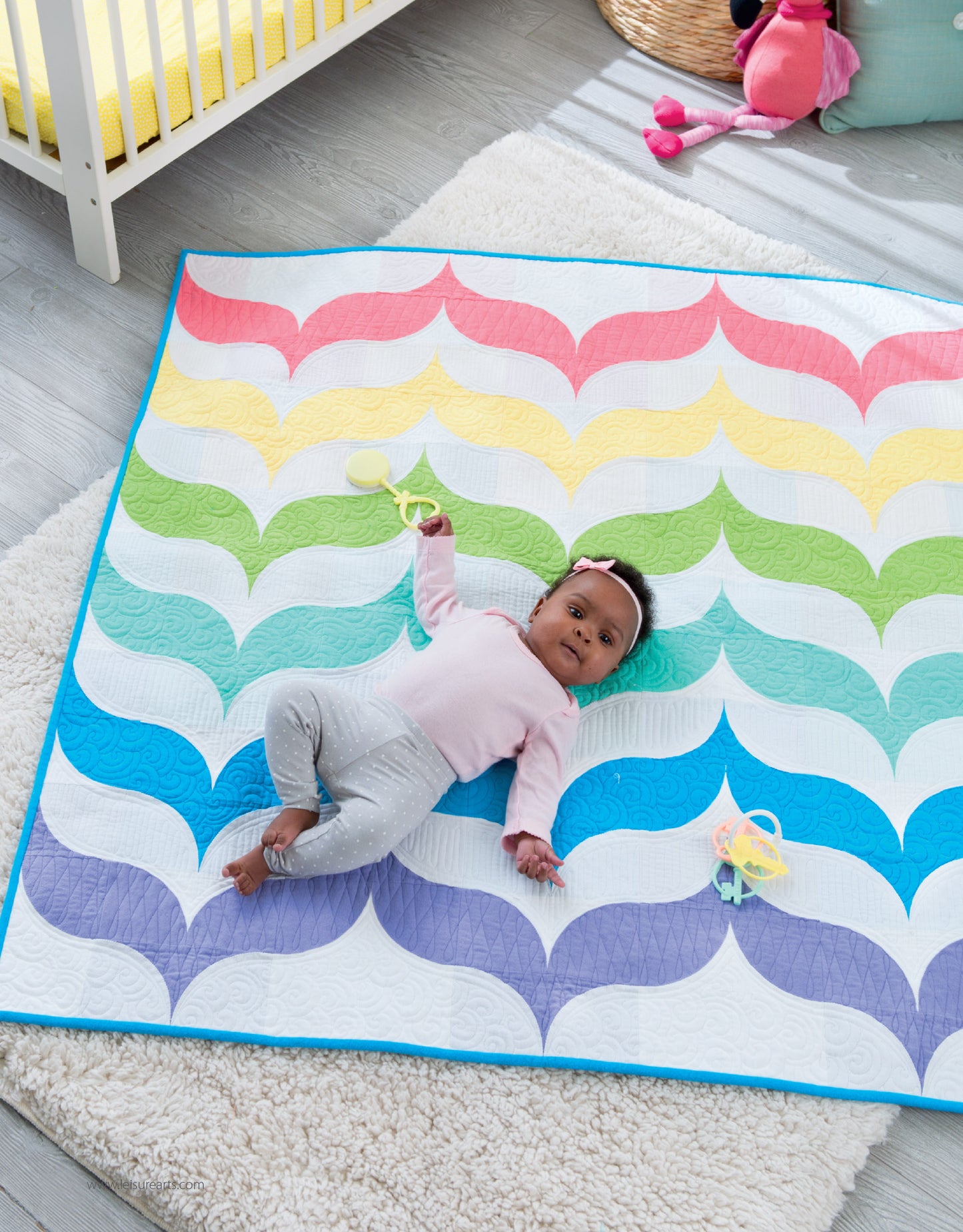 A baby relaxes on a handmade quilt from Leisure Arts' "Baby Quilts—7 Modern Designs to Make Great Gifts Plus Bonus Online Projects," featuring rainbow scallops. Nearby are toys and a crib, and the baby wears a pink top, gray pants, and a white headband.