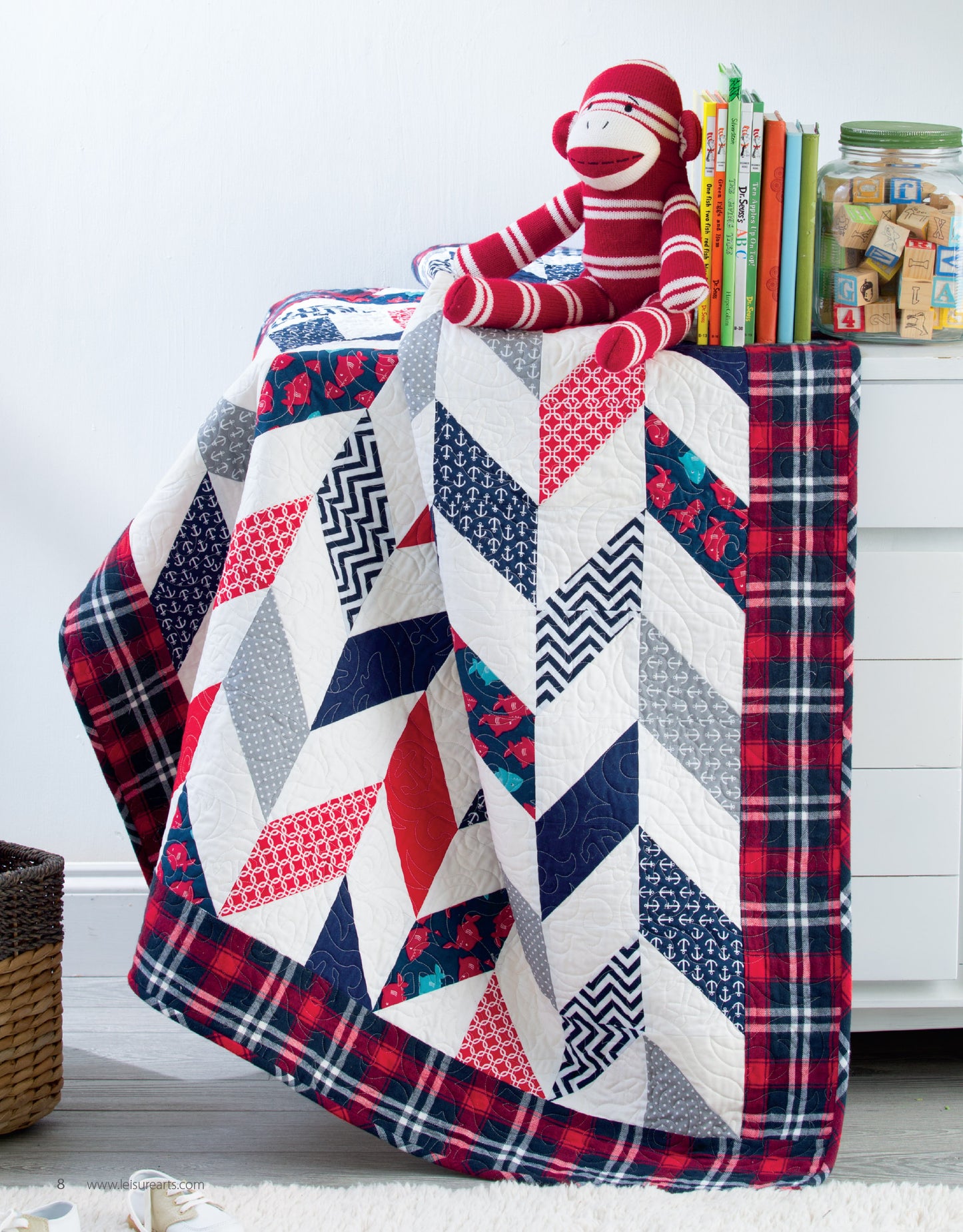 The "Baby Quilts—7 Modern Designs to Make Great Gifts Plus Bonus Online Projects" book by Leisure Arts sits atop a dresser with a red sock monkey, children's books, and a jar of wooden blocks, displaying charming quilt patterns.