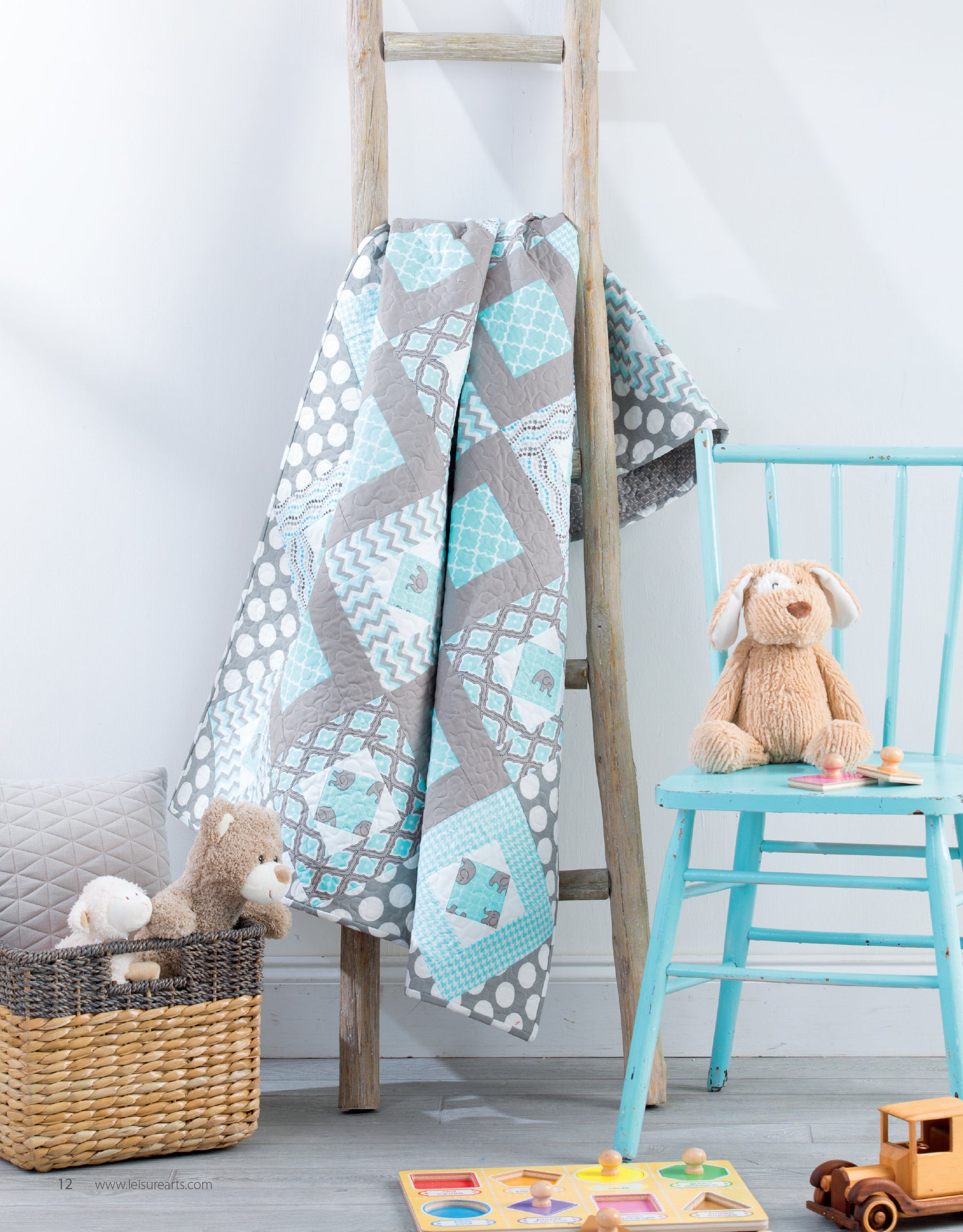 A handmade quilt from Leisure Arts’ “Baby Quilts—7 Modern Designs to Make Great Gifts Plus Bonus Online Projects” hangs on a rustic ladder beside a light blue chair with a plush bunny, with toys and puzzles arranged nearby.