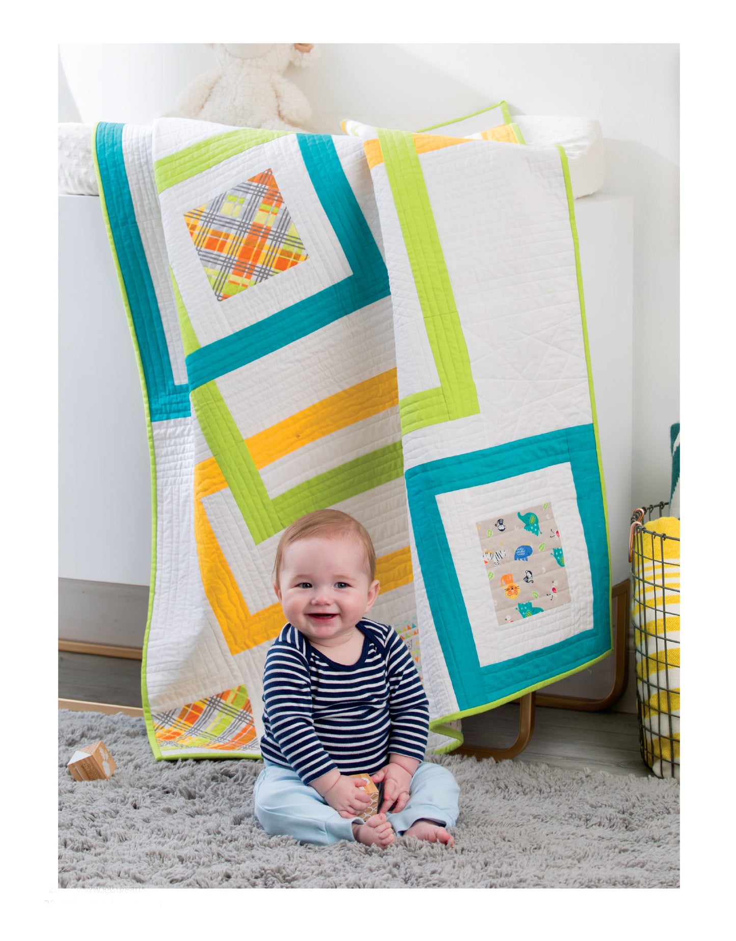 A smiling baby in a striped shirt and blue pants sits before a crib draped with a colorful quilt made from Leisure Arts' "Baby Quilts—7 Modern Designs to Make Great Gifts Plus Bonus Online Projects." A stuffed animal peeks out behind the vibrant patterns.