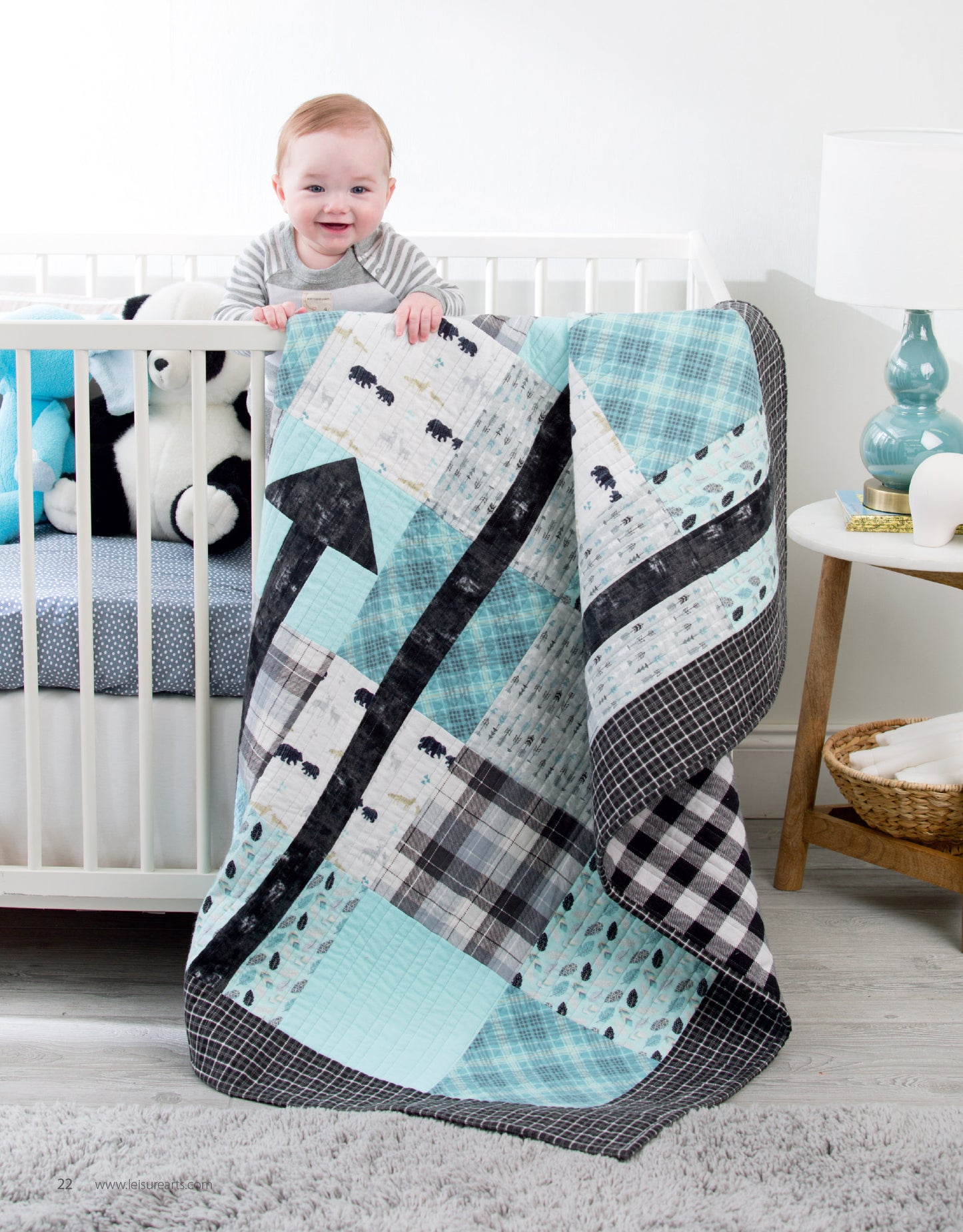 A smiling baby stands in a white crib, holding a blue, black, and white patchwork quilt made using “Baby Quilts—7 Modern Designs to Make Great Gifts Plus Bonus Online Projects” from Leisure Arts; a stuffed panda and polka-dot bedding complete the cozy scene.