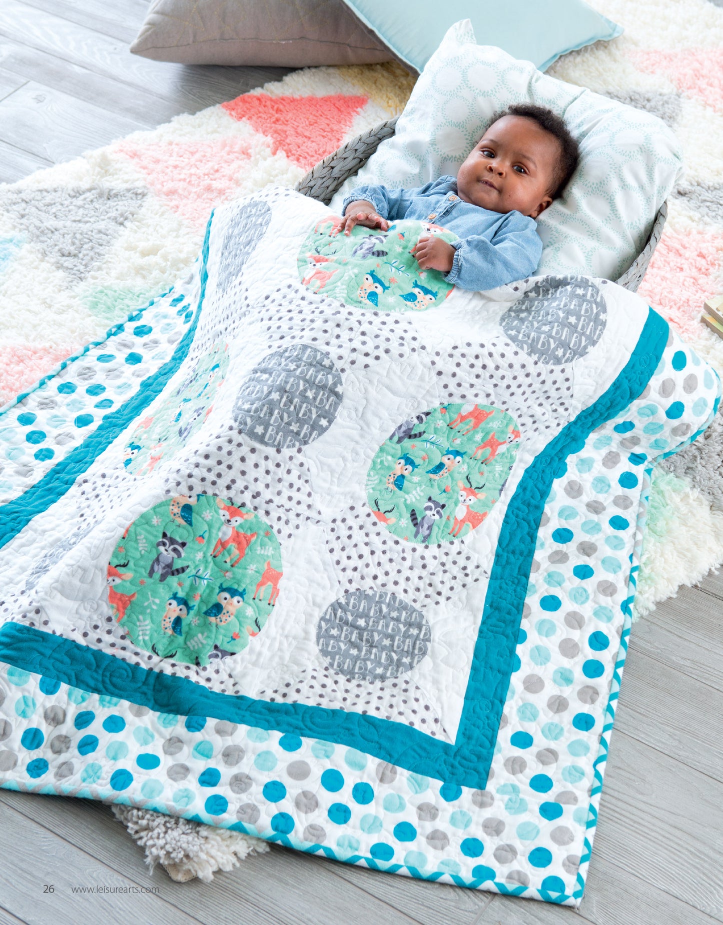 A baby smiles and rests on a patterned pillow, covered with a colorful handmade quilt from "Baby Quilts—7 Modern Designs to Make Great Gifts Plus Bonus Online Projects" by Leisure Arts. The cozy scene features teal borders and circular patterns on the soft rug.
