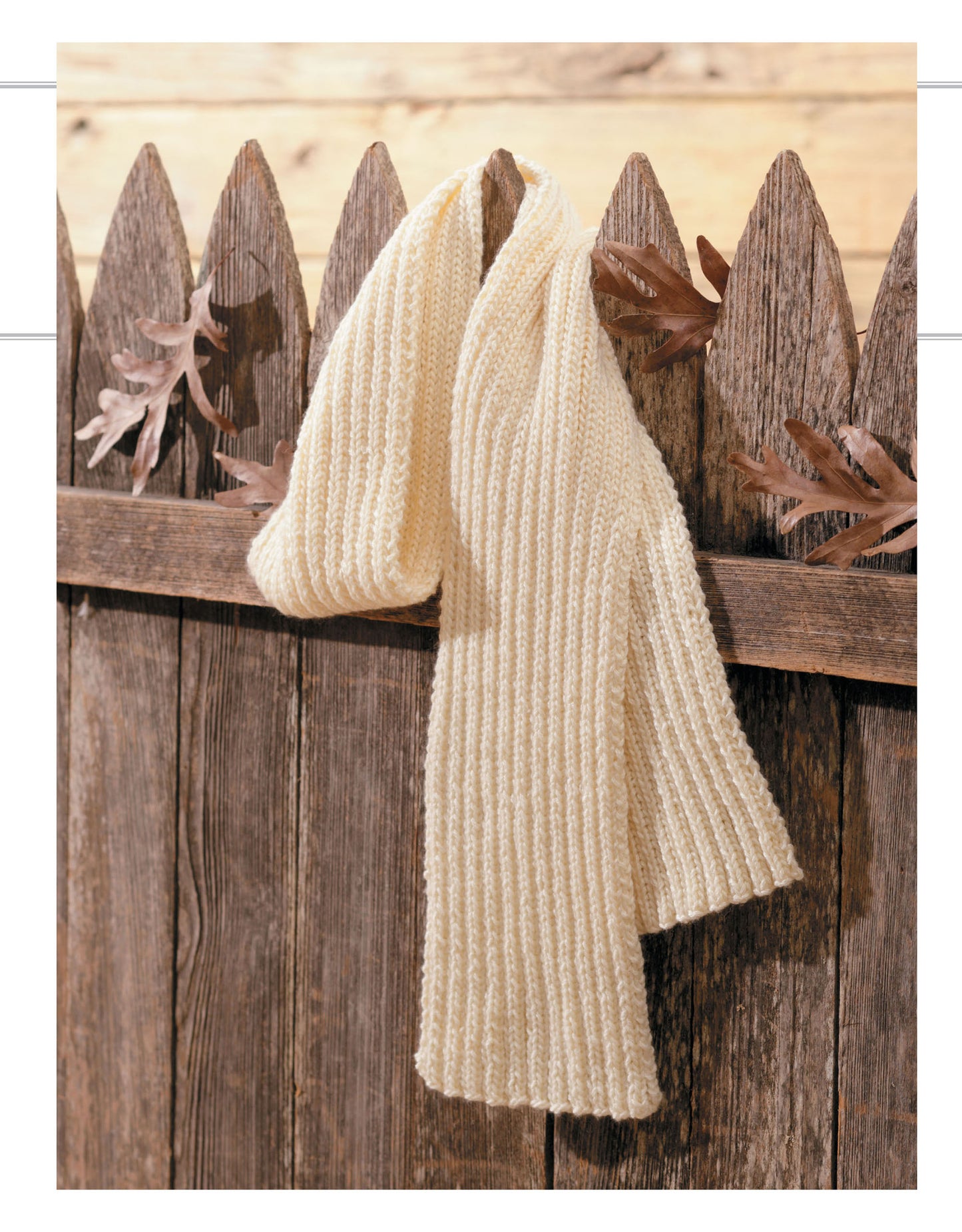 A cream Cozy Scarves Knitting scarf by Leisure Arts, featuring easy rib knit instructions, drapes over a rustic wooden picket fence with scattered oak leaves. The scene is set against pale wooden boards.