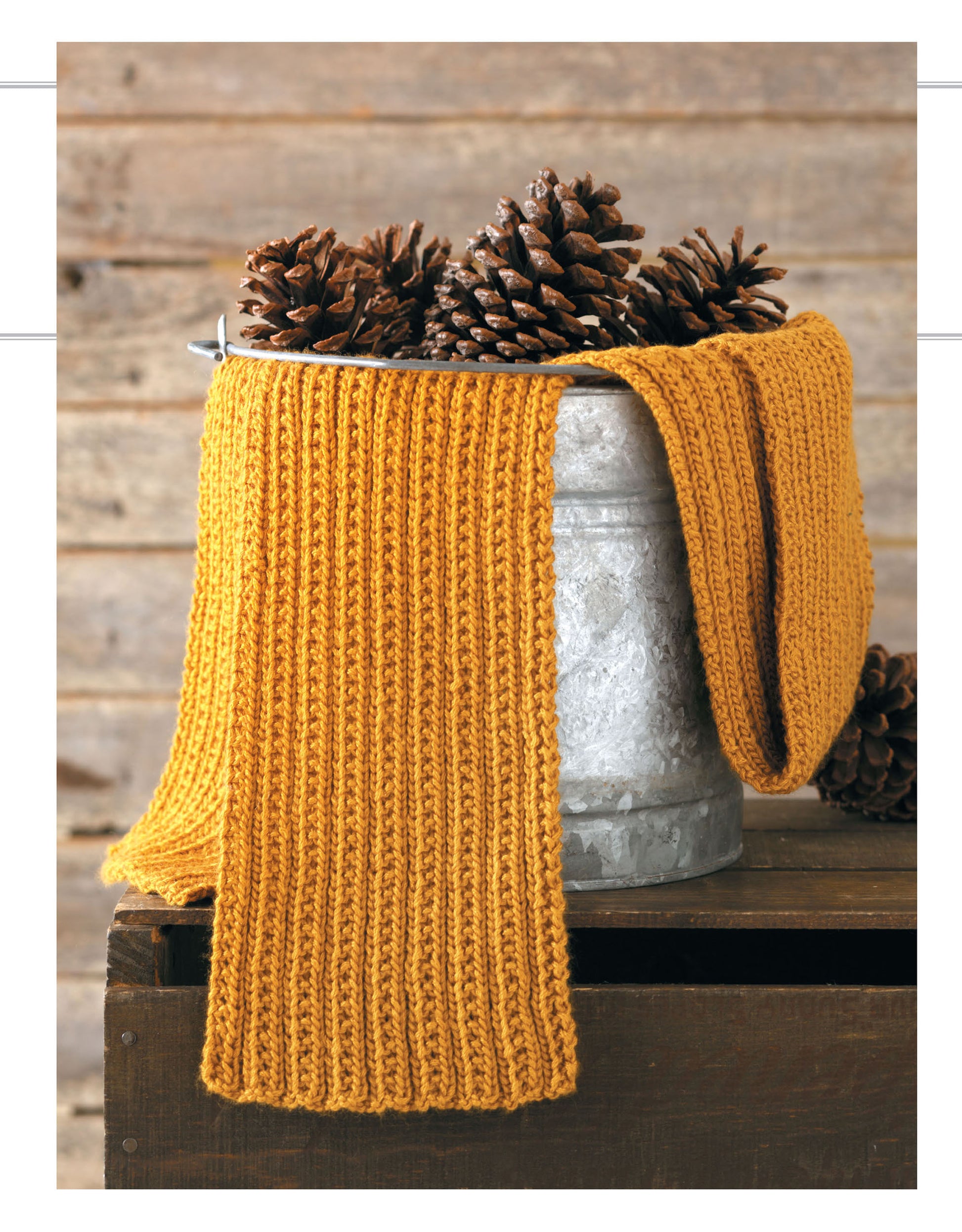 A Leisure Arts Cozy Scarves Knitting ribbed mustard yellow scarf is draped over a pine cone-filled bucket on rustic wood, with a wooden plank wall backdrop—ideal for beginner knitters.
