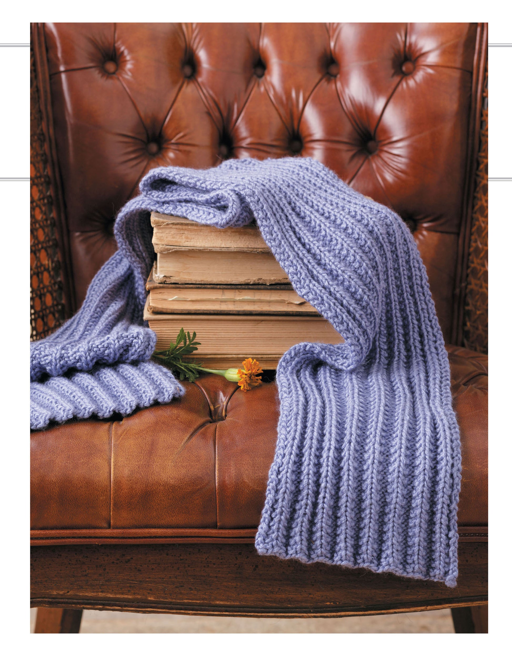 A stack of old books sits on a tufted leather chair, topped with a chunky lavender scarf from Leisure Arts’ Cozy Scarves Knitting—ideal for beginners. A small orange flower and green leaf add charm to the scene.