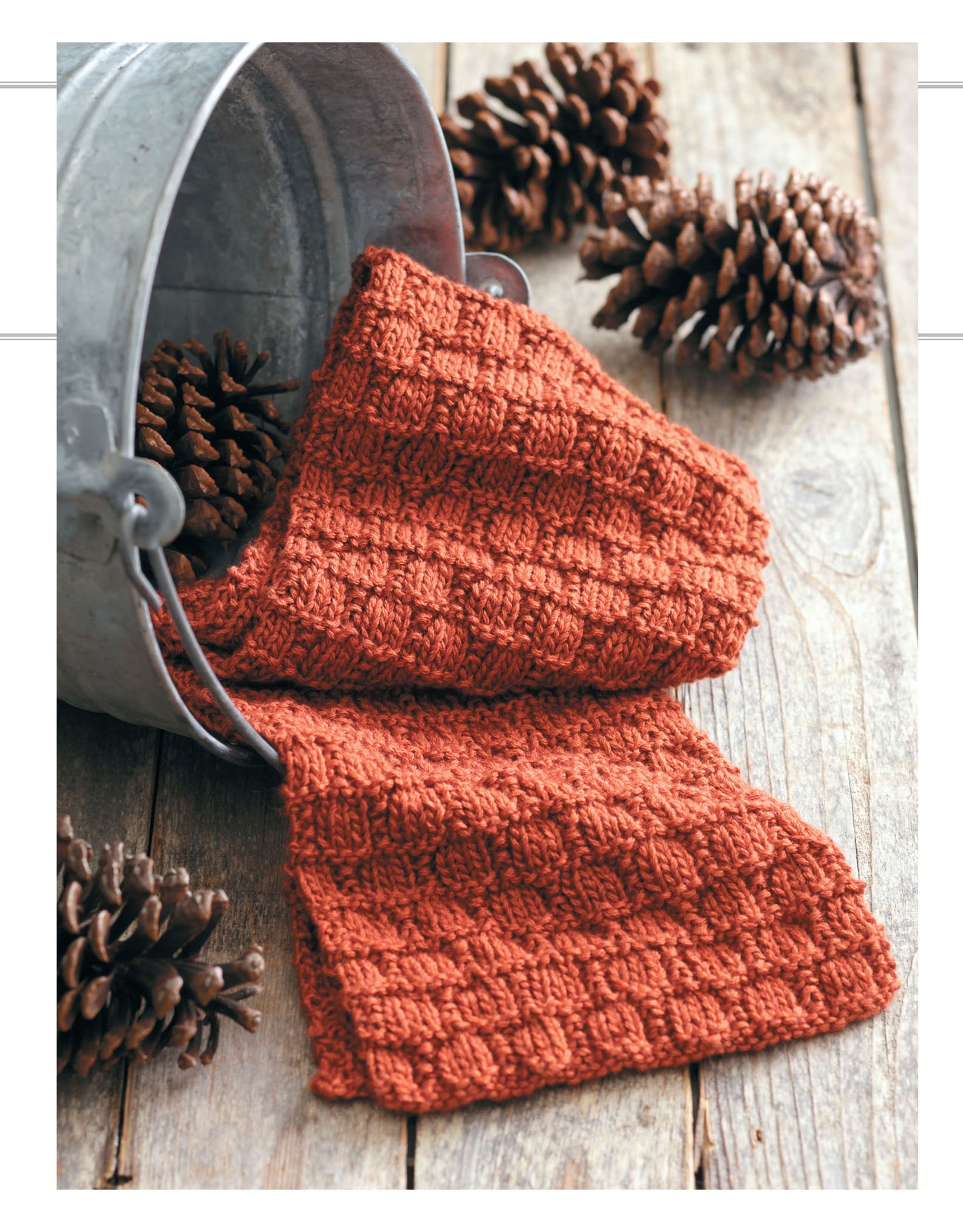 A rust-colored rib knit scarf from Leisure Arts' Cozy Scarves Knitting drapes from a metal bucket onto rustic wood, surrounded by pinecones—an ideal beginner knitting project.