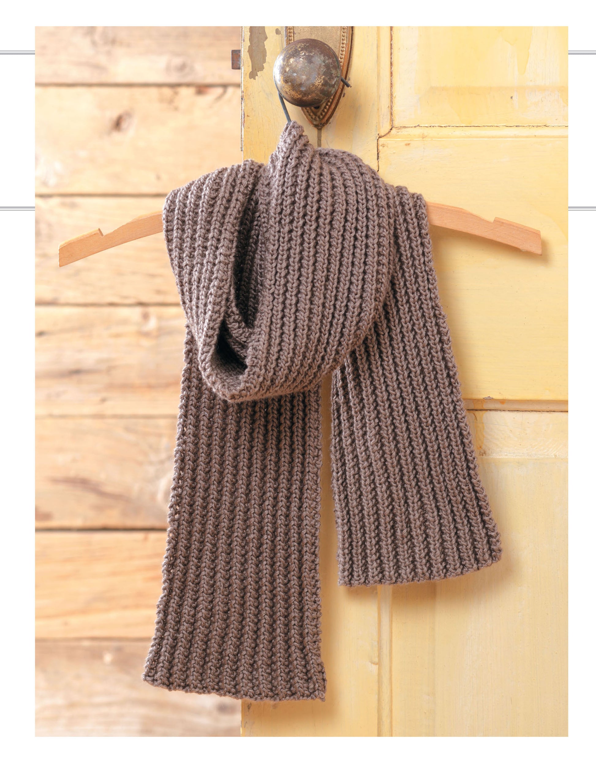 The Cozy Scarves Knitting scarf by Leisure Arts, a thick ribbed brown knit perfect for beginners, is draped on a wooden hanger hung from a round metal knob on a yellow door with light wood plank background.